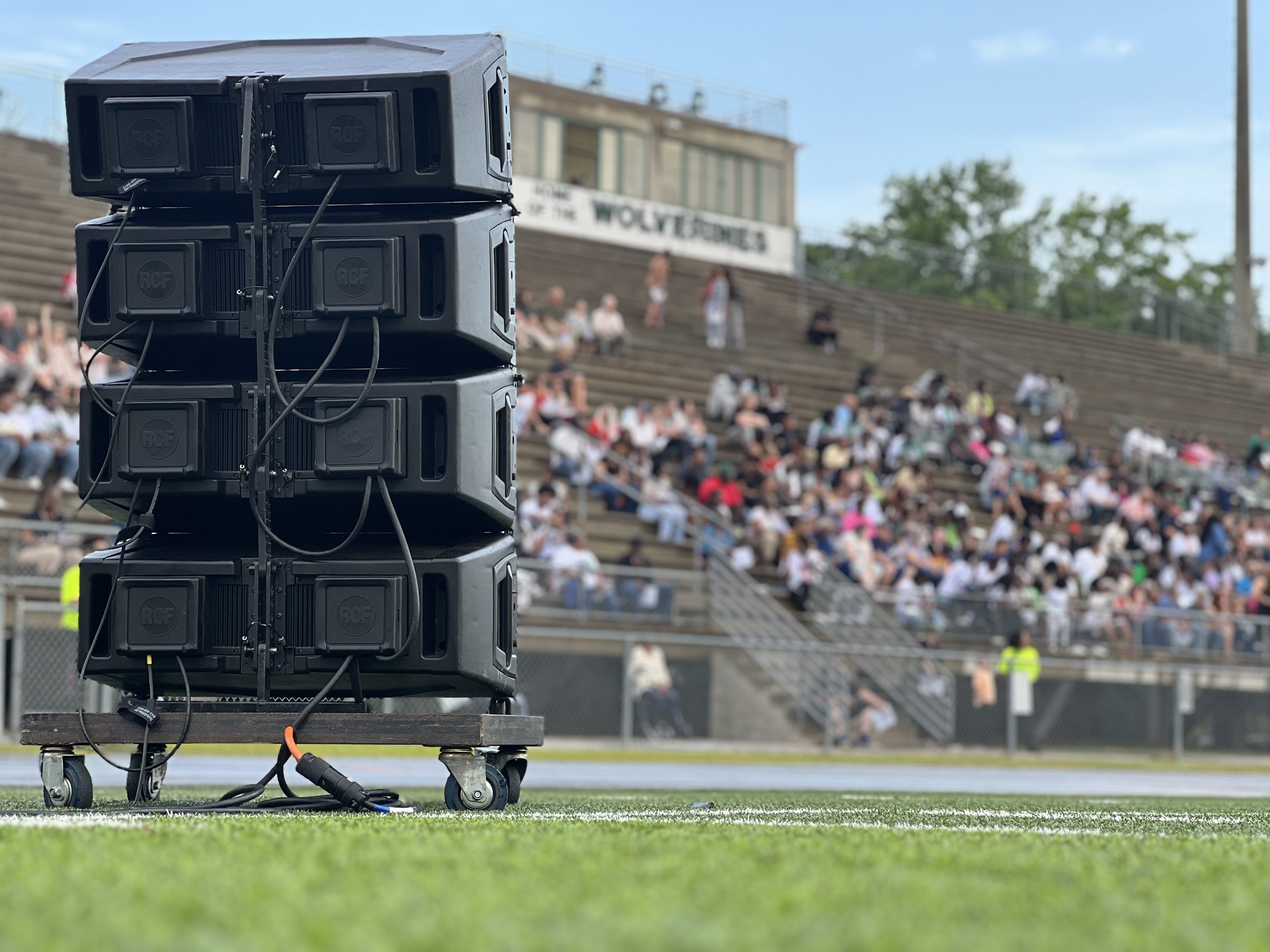 Large black speakers on a stage on a sports field, with a blurred crowd sitting on bleachers in the background.