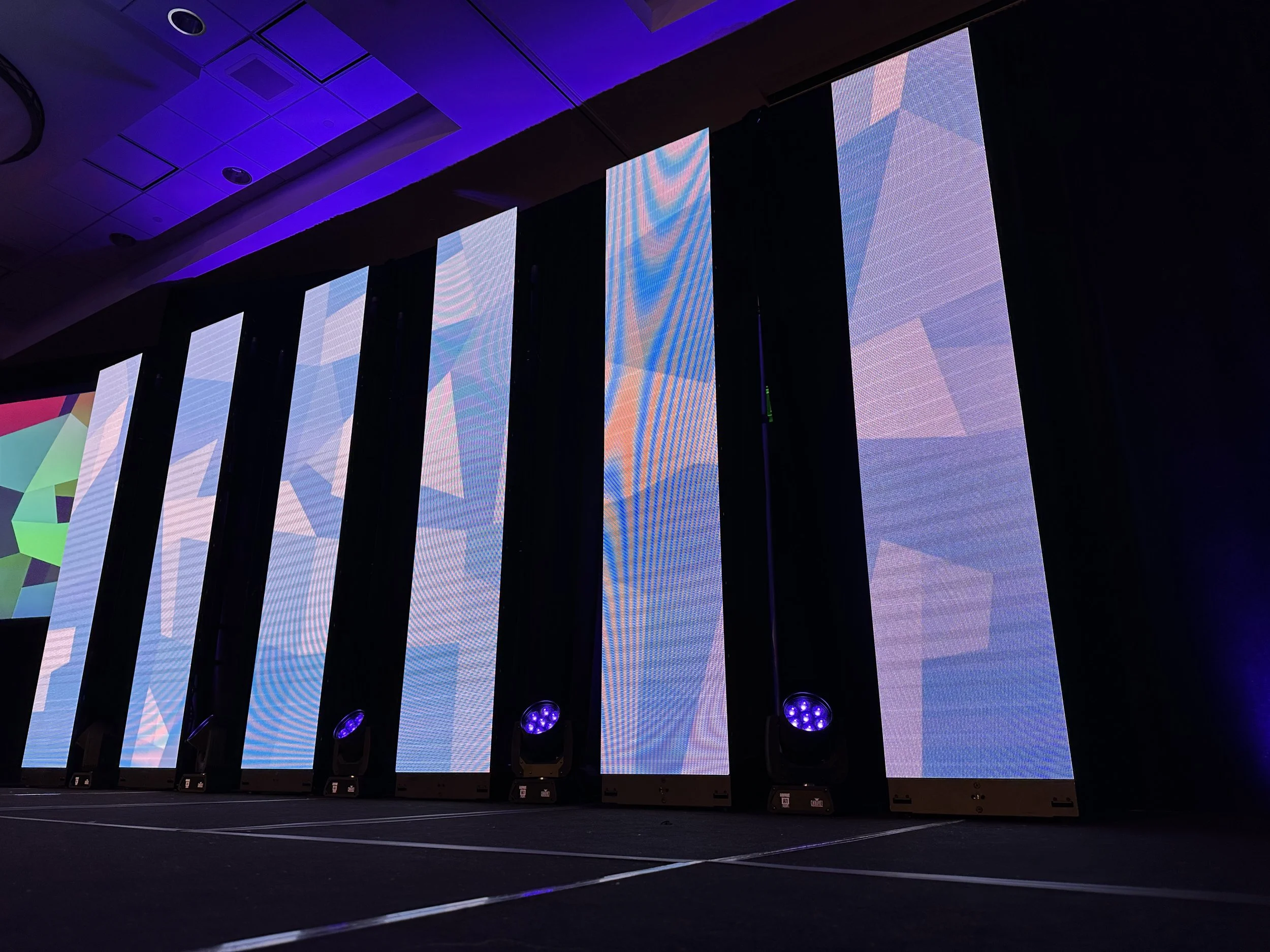 Multiple large vertical LED screens displaying colorful abstract patterns in a darkened conference room or stage setting.
