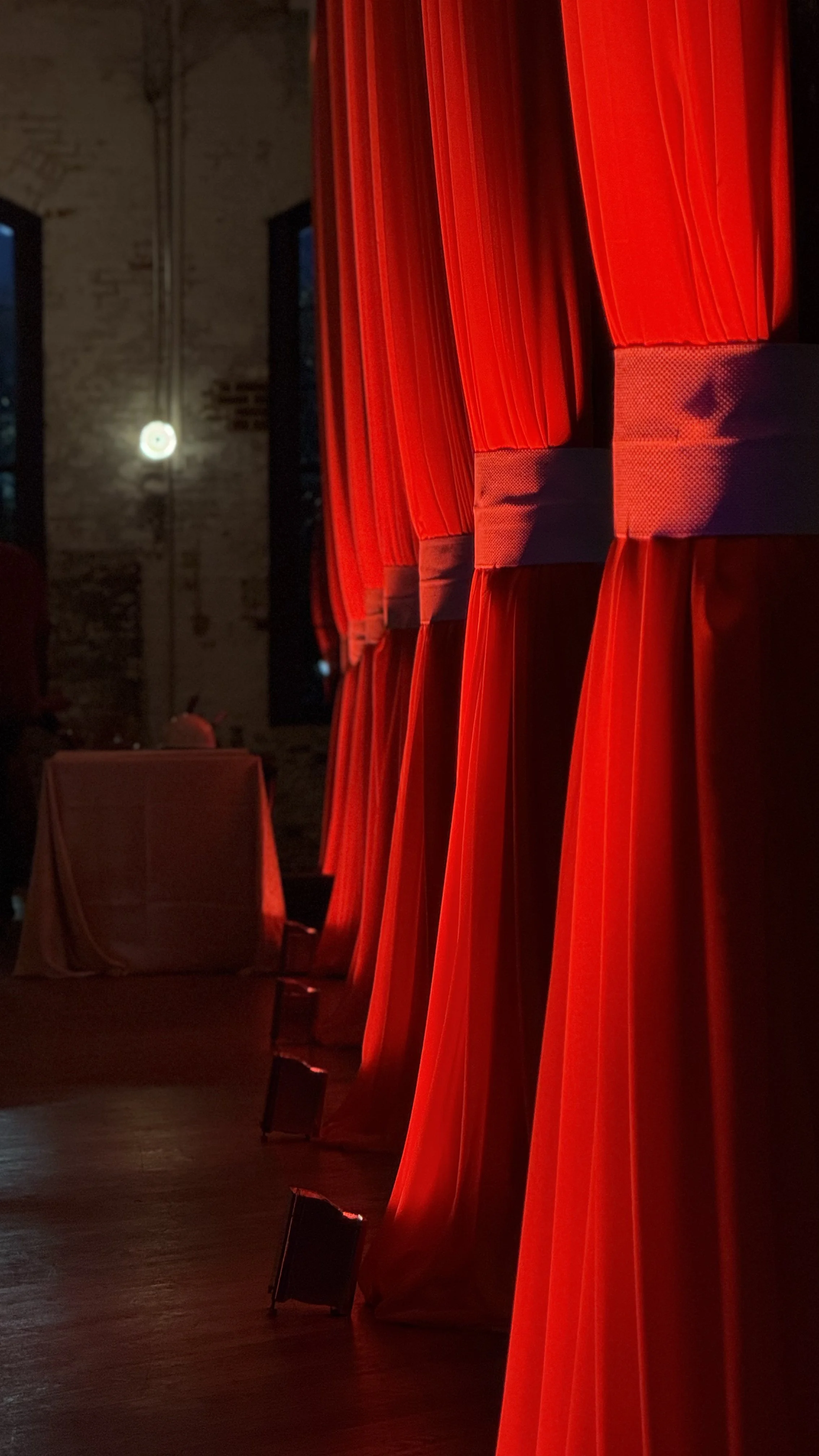 Row of red theater curtains with a stool and table in the background, brick wall interior with a hanging light.