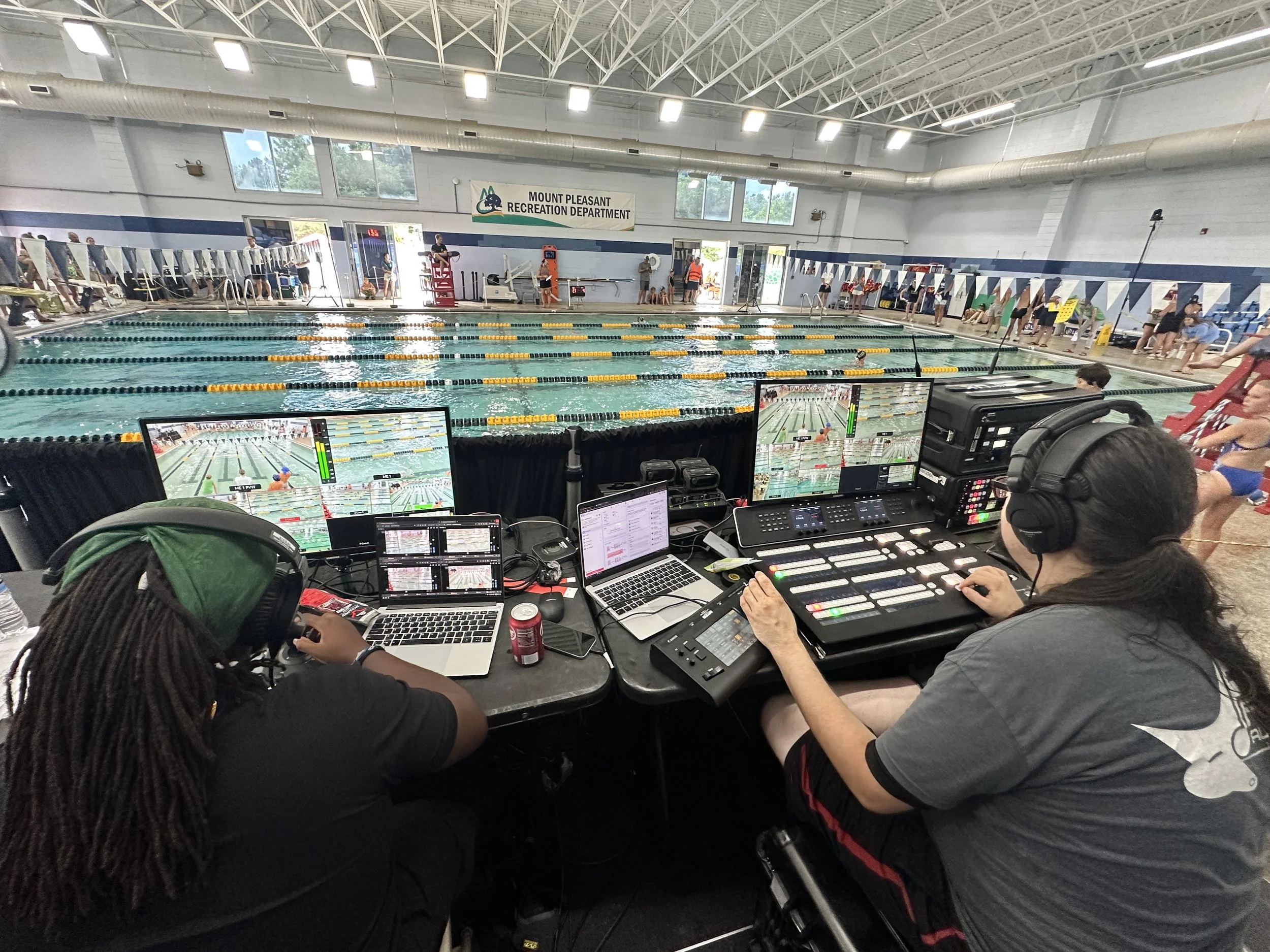 Indoor swimming pool with people lined up along the edge and a swimmer in the pool. Control panels, laptops, and audio equipment are in the foreground, indicating a live broadcast or event coverage.