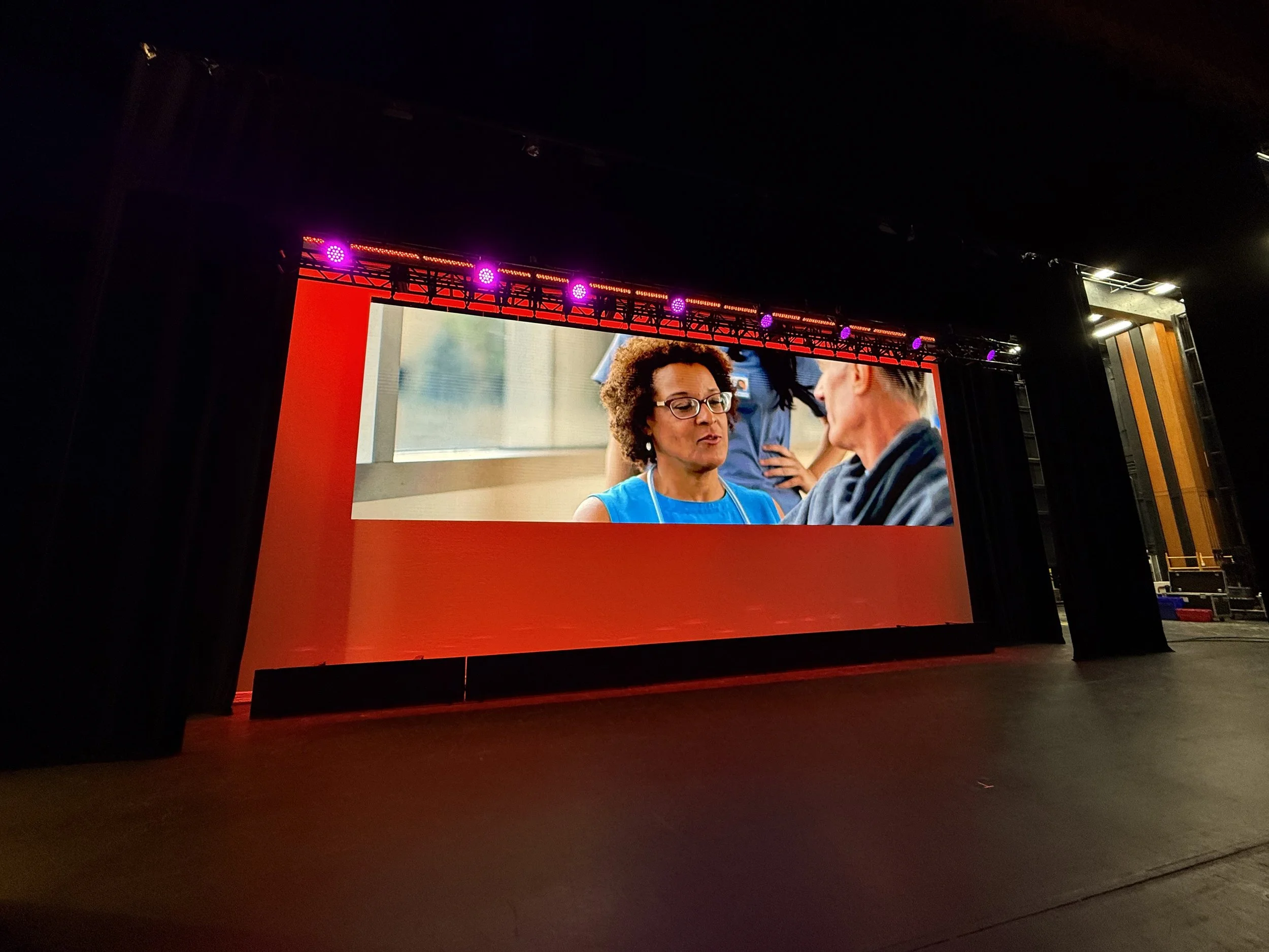 Large indoor stage with a big screen showing two women in conversation, one wearing glasses and a blue top, the other with short gray hair, in a well-lit, colorful setting.