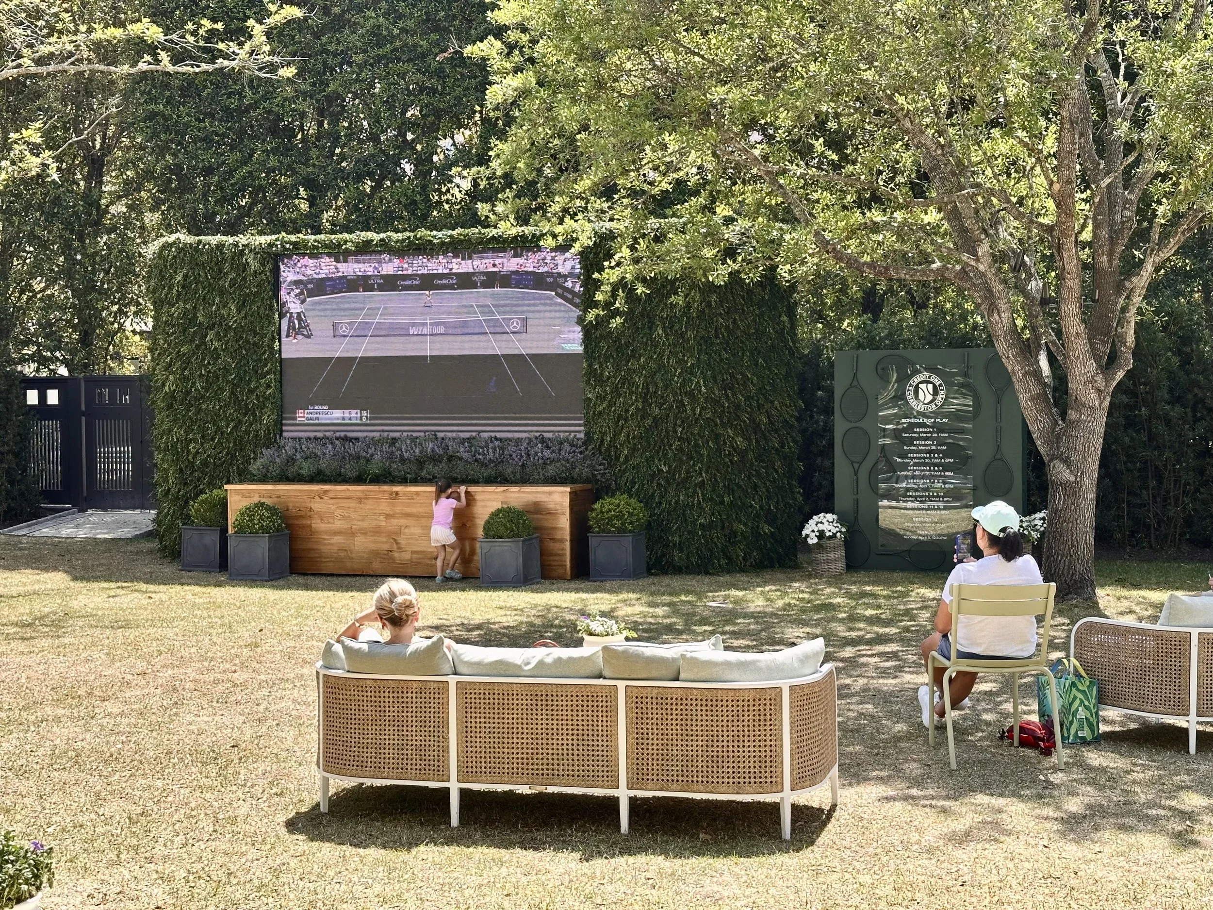 People sitting outdoors in a garden watching a tennis match on a large screen, with trees and greenery surrounding the area.