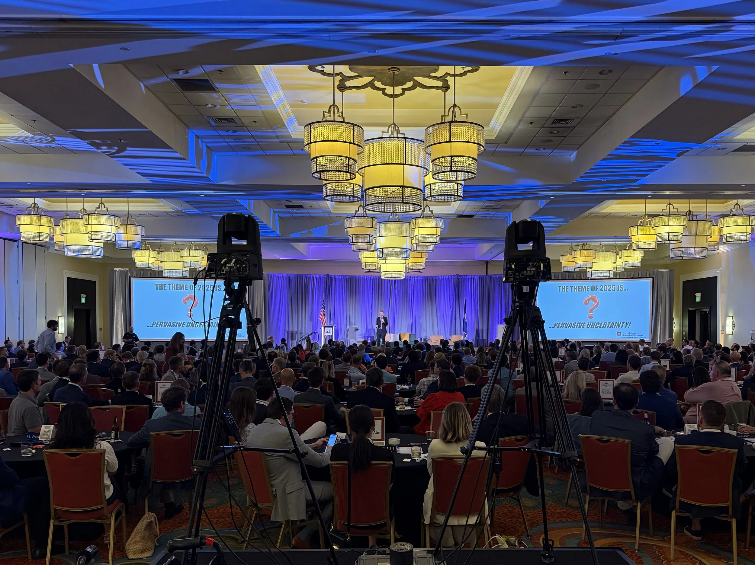 Conference room with a large audience facing a stage with a speaker, two screens displaying a presentation, and ceiling lights.