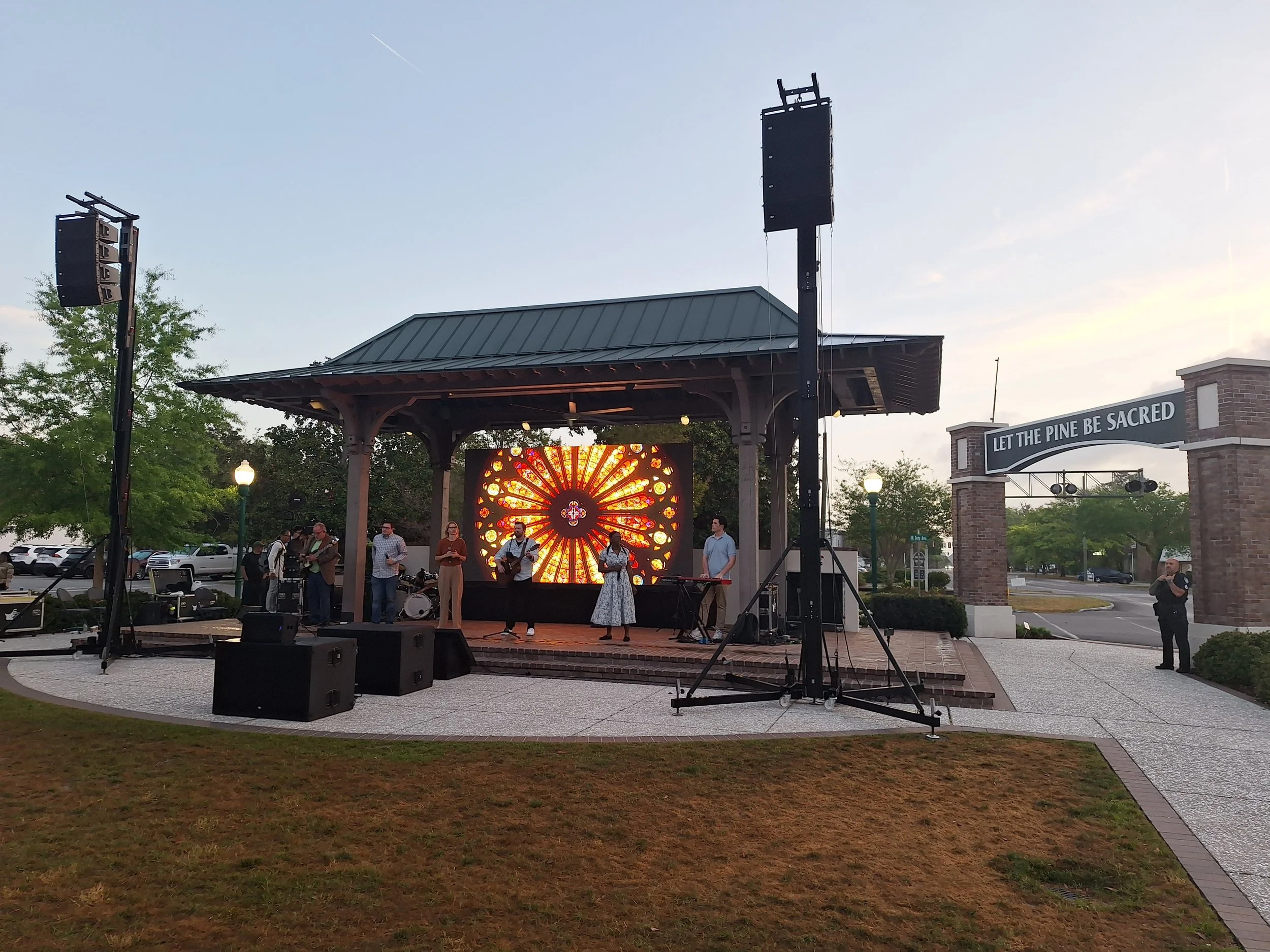 A small outdoor stage with musicians performing, including a woman singing and playing guitar, a man playing guitar, and other band members. Behind them is a digital screen displaying a stained glass window design. The stage is in a park with trees a