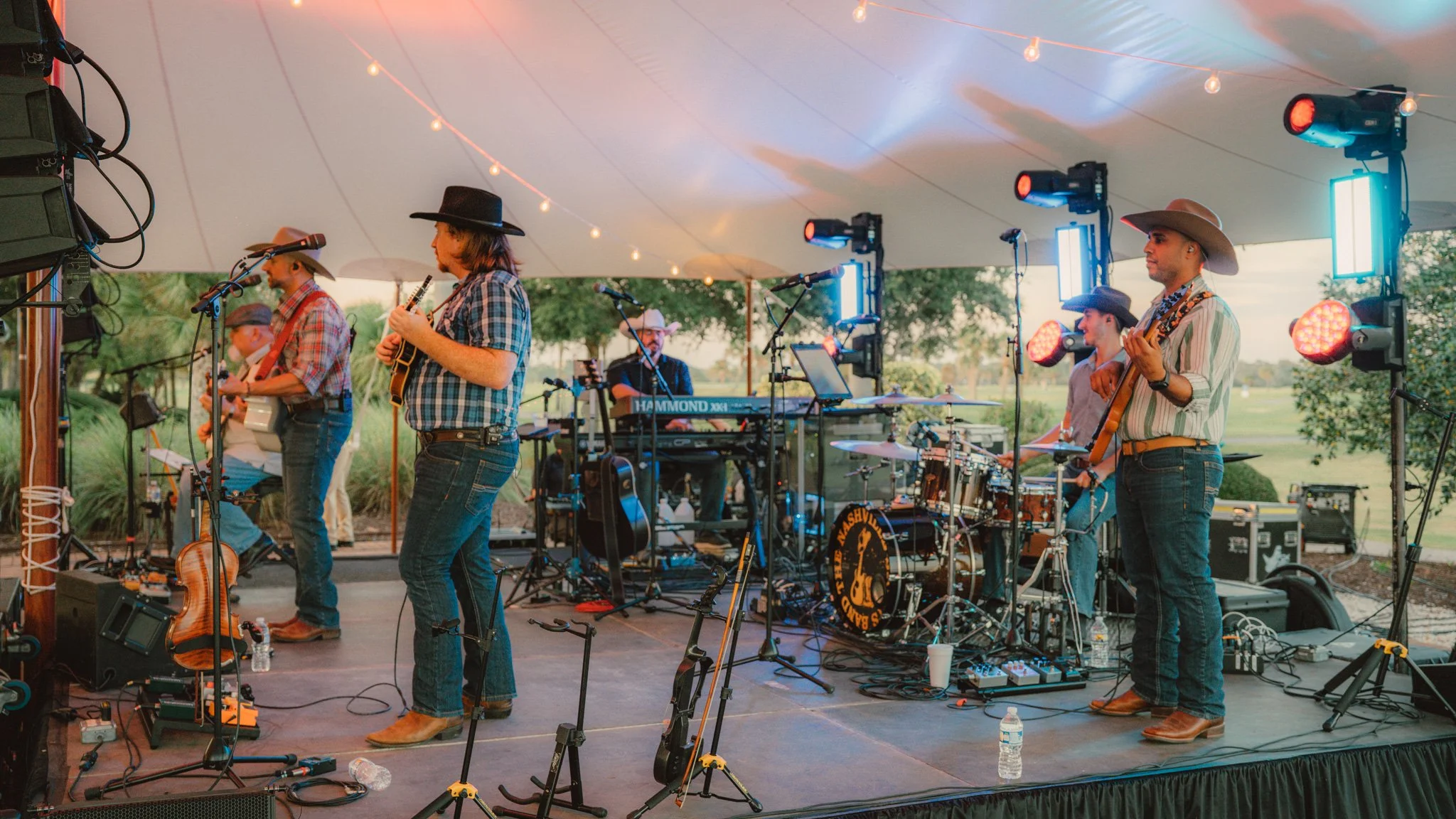 A band performing on an outdoor stage under a tent with string lights, with musicians playing guitars, drums, keyboard, and singing, wearing cowboy hats and casual country attire.