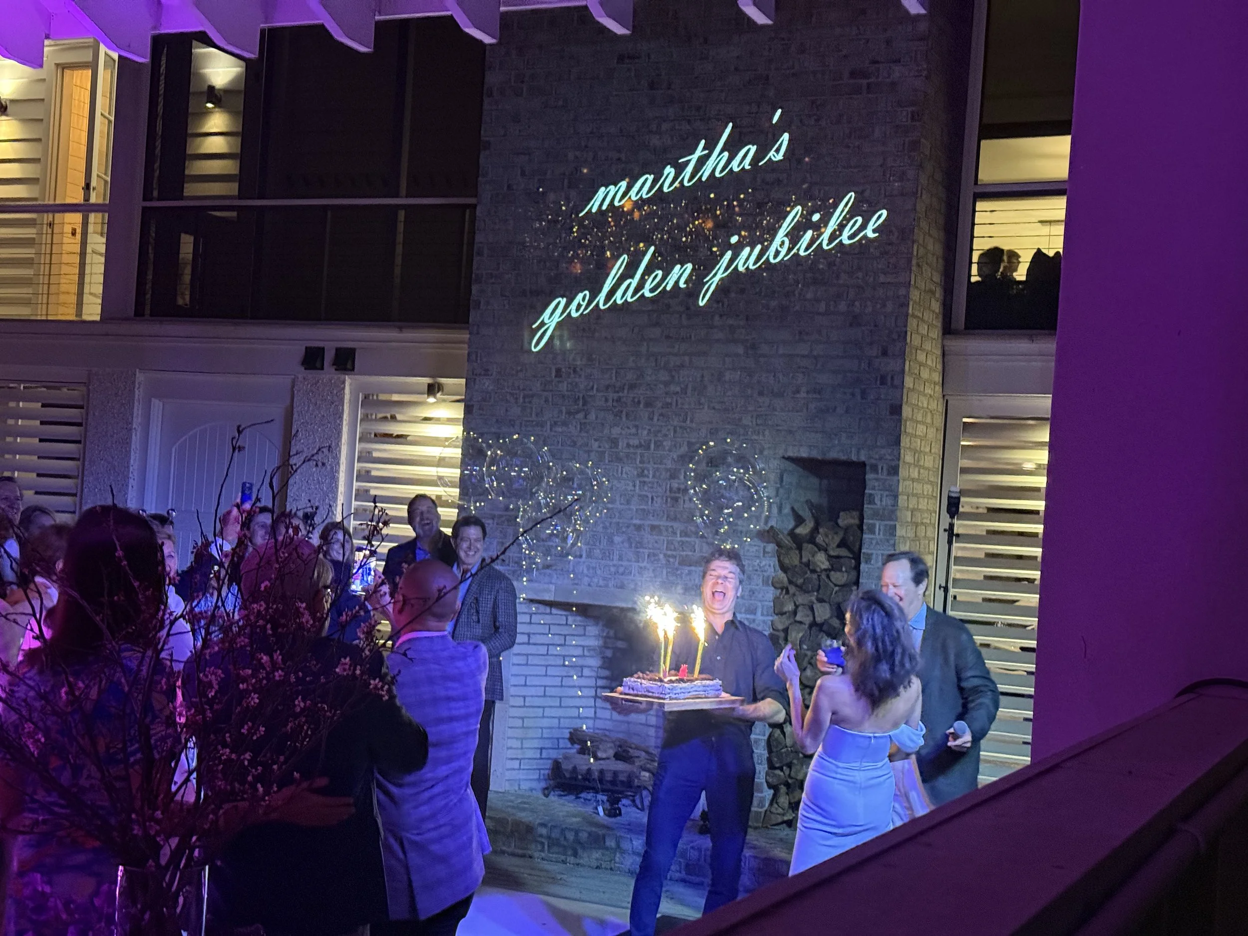 A birthday celebration with people gathered outdoors at night, with a woman holding a cake with sparklers. The wall behind has a sign that reads 'martha's golden jubilee'.