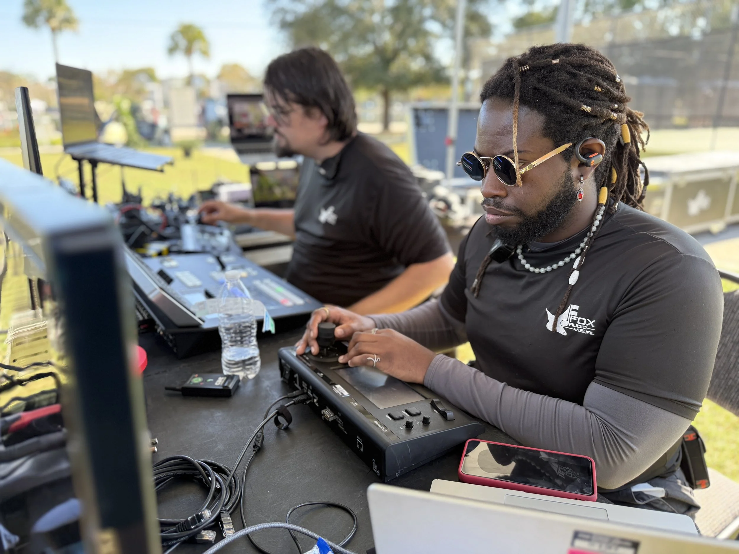 Two DJs are working at an outdoor event. The DJ in the foreground has dreadlocks, sunglasses, and is using a DJ mixer. The second DJ in the background is focused on his equipment. There are electronic devices, water bottles, and a smartphone on the t