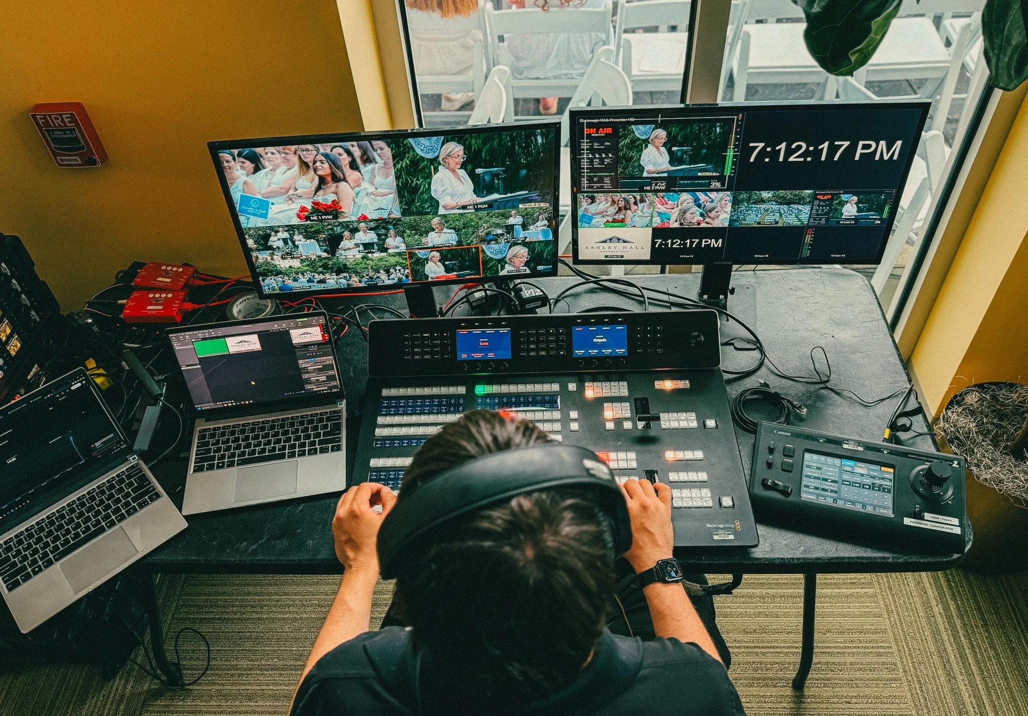 A control room with multiple monitors showing a live event of a woman speaking at an outdoor event, with a crowd and greenery in the background. The person operating the equipment is wearing headphones and a wristwatch, sitting at a desk with various