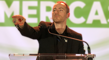 A man in a black suit pointing forward while speaking at a podium during a presentation or speech, with a green and white background that appears to have the word 'MERCA'.