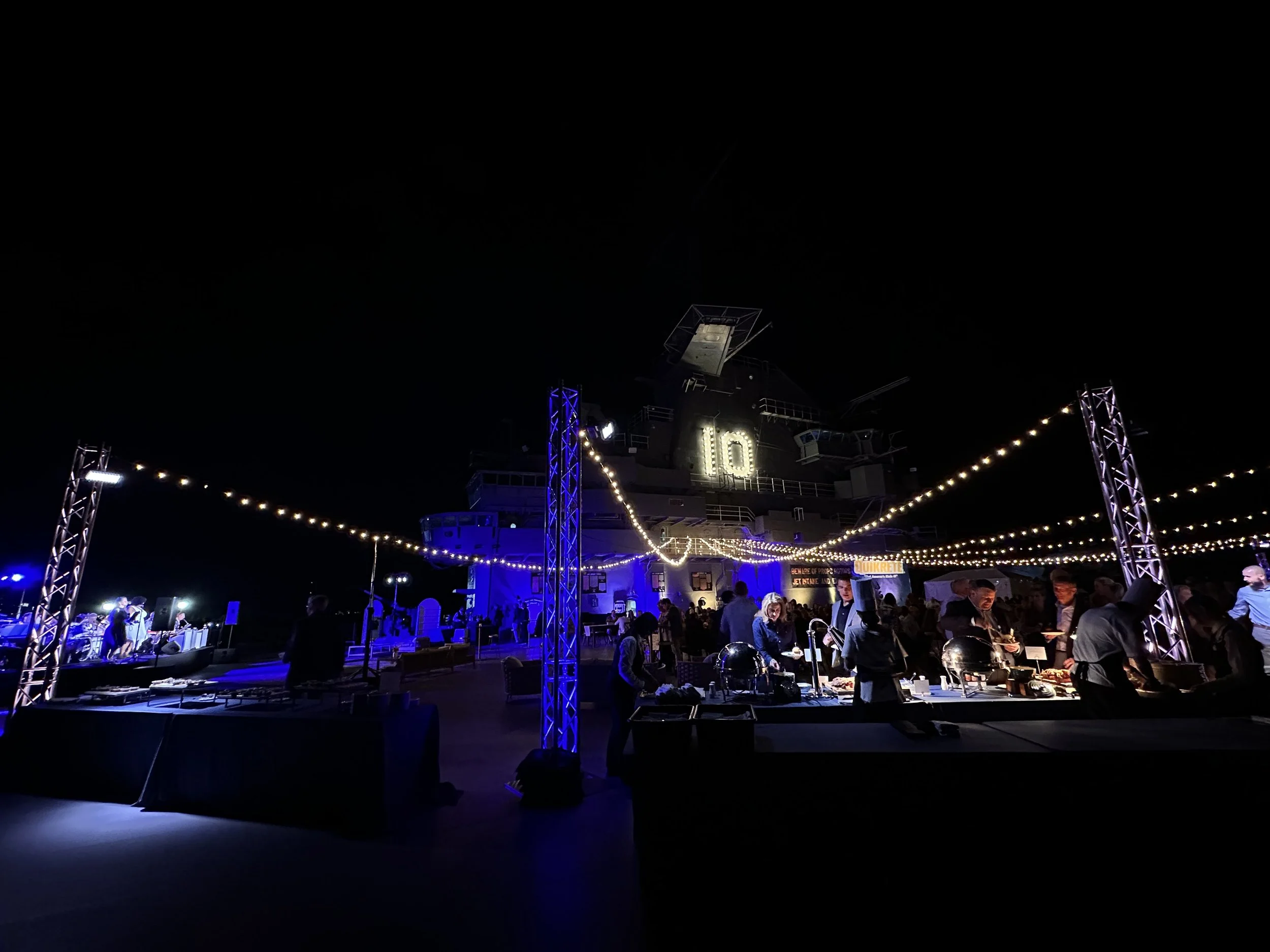 Nighttime outdoor event on a ship deck with string lights, people serving food at a buffet, and a large illuminated number 10 on the ship's superstructure.