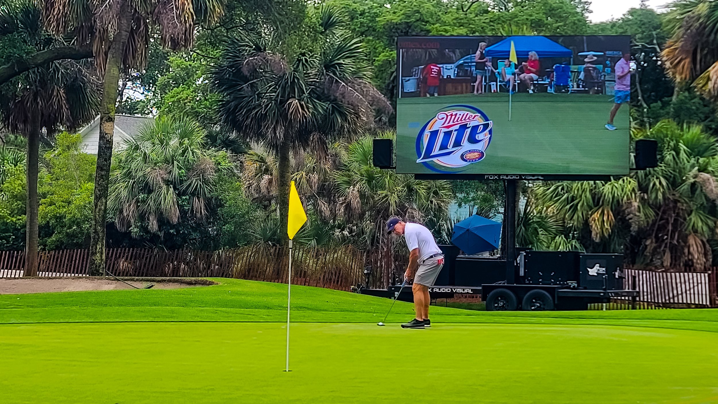 A man is preparing to hit a golf ball on a golf course, with a large TV screen showing a live feed of the hole and nearby spectators in the background.