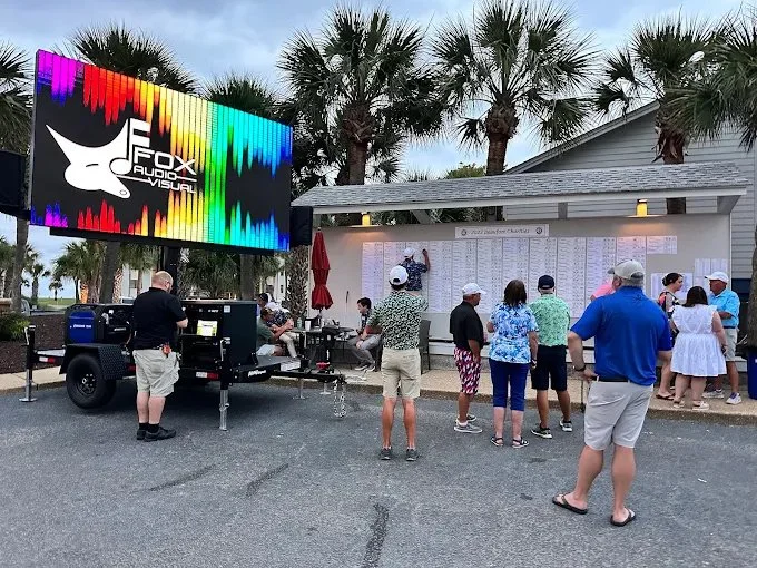Groups of people looking at a large board with scores outside, next to a digital screen displaying a colorful audio wave and the logo 'Fox Audio Visual' at a golf course.