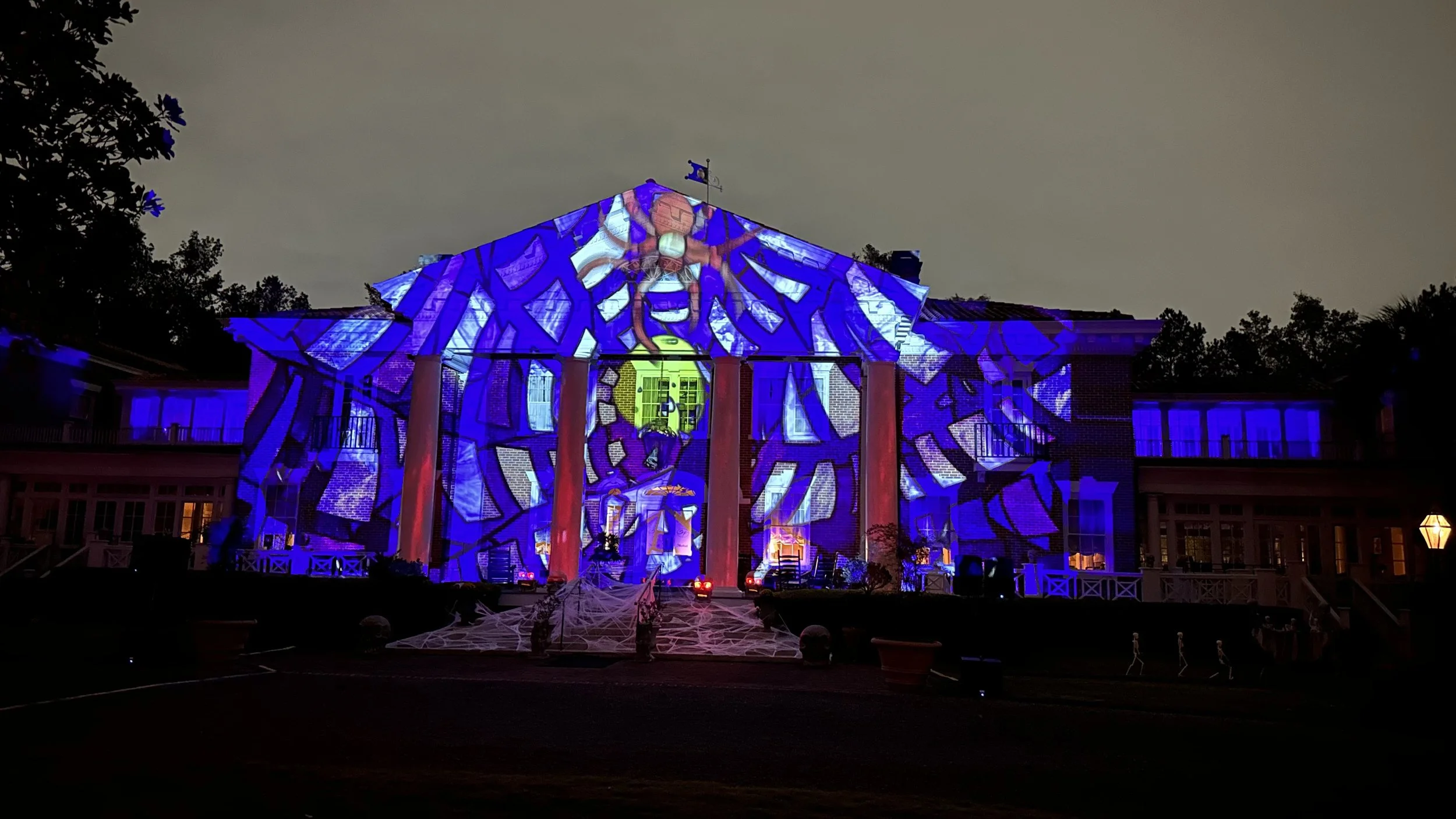 A house decorated with colorful projected lights and images for Halloween, featuring a large spider and web design on the front facade at night.