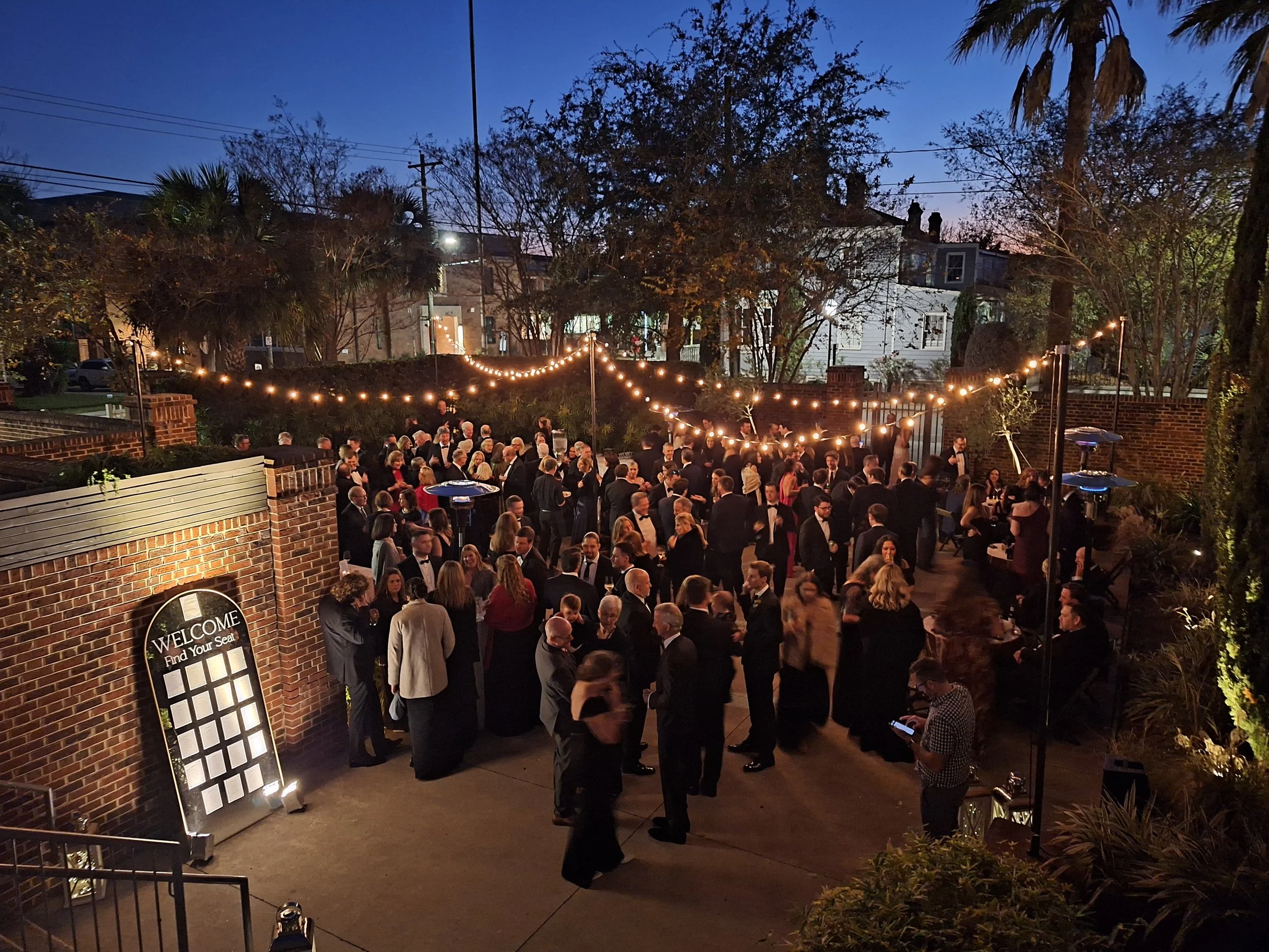 Outdoor evening event with a large crowd of people dressed in formal attire, decorated with string lights, at a venue with brick walls and trees.