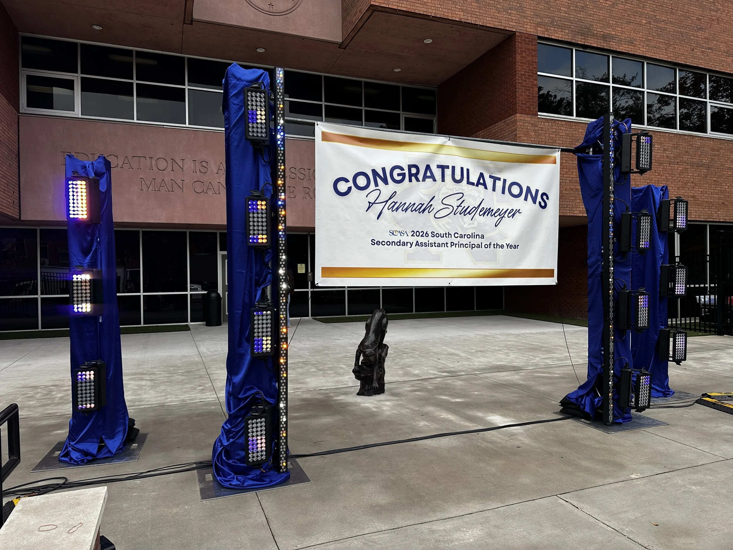 Graduation stage setup with a large congratulatory banner for Hannah Studemeyer, who was named South Carolina Secondary Assistant Principal of the Year 2026, outside a modern brick building. The stage features tall blue-covered structures with lighti