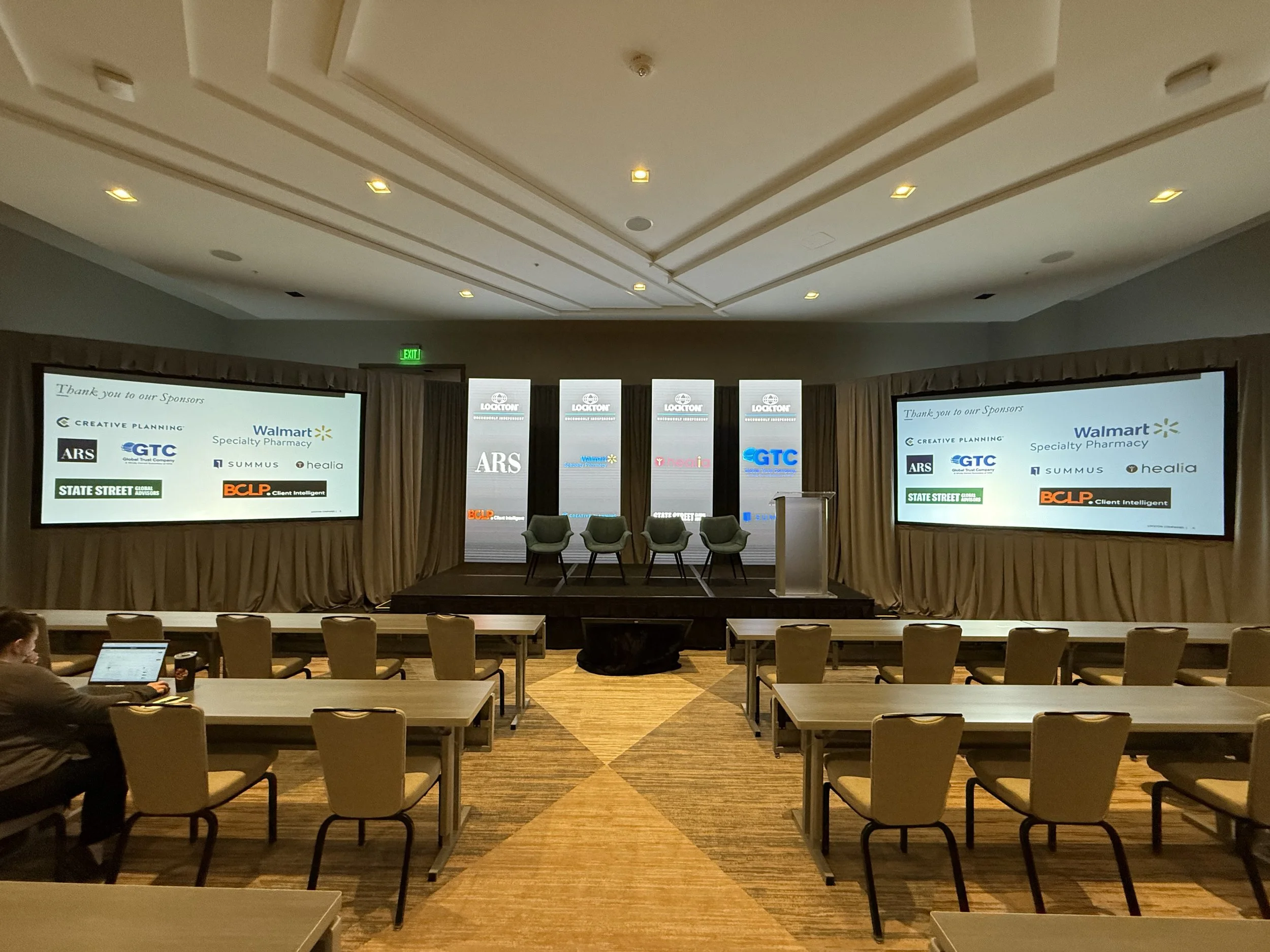 Conference room set up with chairs and tables, two large screens displaying sponsor logos, and a stage with four chairs and a podium.