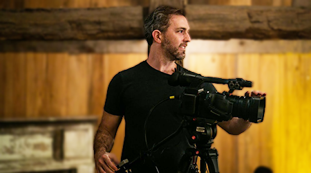 A man with a beard operating a professional video camera in a wood-paneled room.