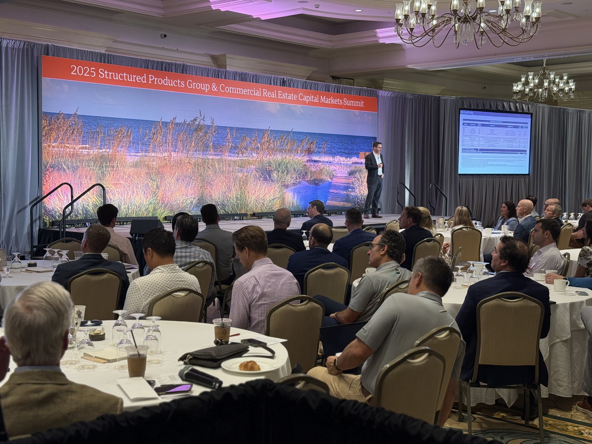 Conference room with attendees watching a speaker on stage. Large screen displays presentation slides. Red banner above reads '2025 Structured Products Group & Commercial Real Estate Capital Markets Summit.' Elegant chandeliers hang from the ceiling,
