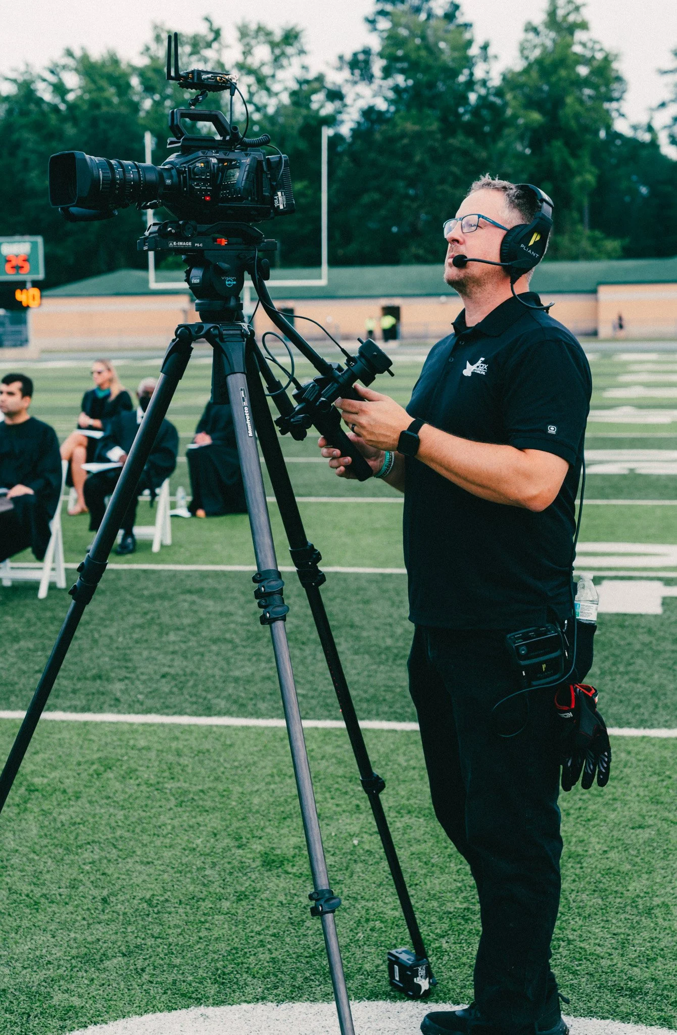 A man wearing glasses and a black polo shirt with a logo operates a large professional video camera on a tripod at a football field during sunset or early evening. The man is wearing a headset with a microphone, and there are seated people in the bac