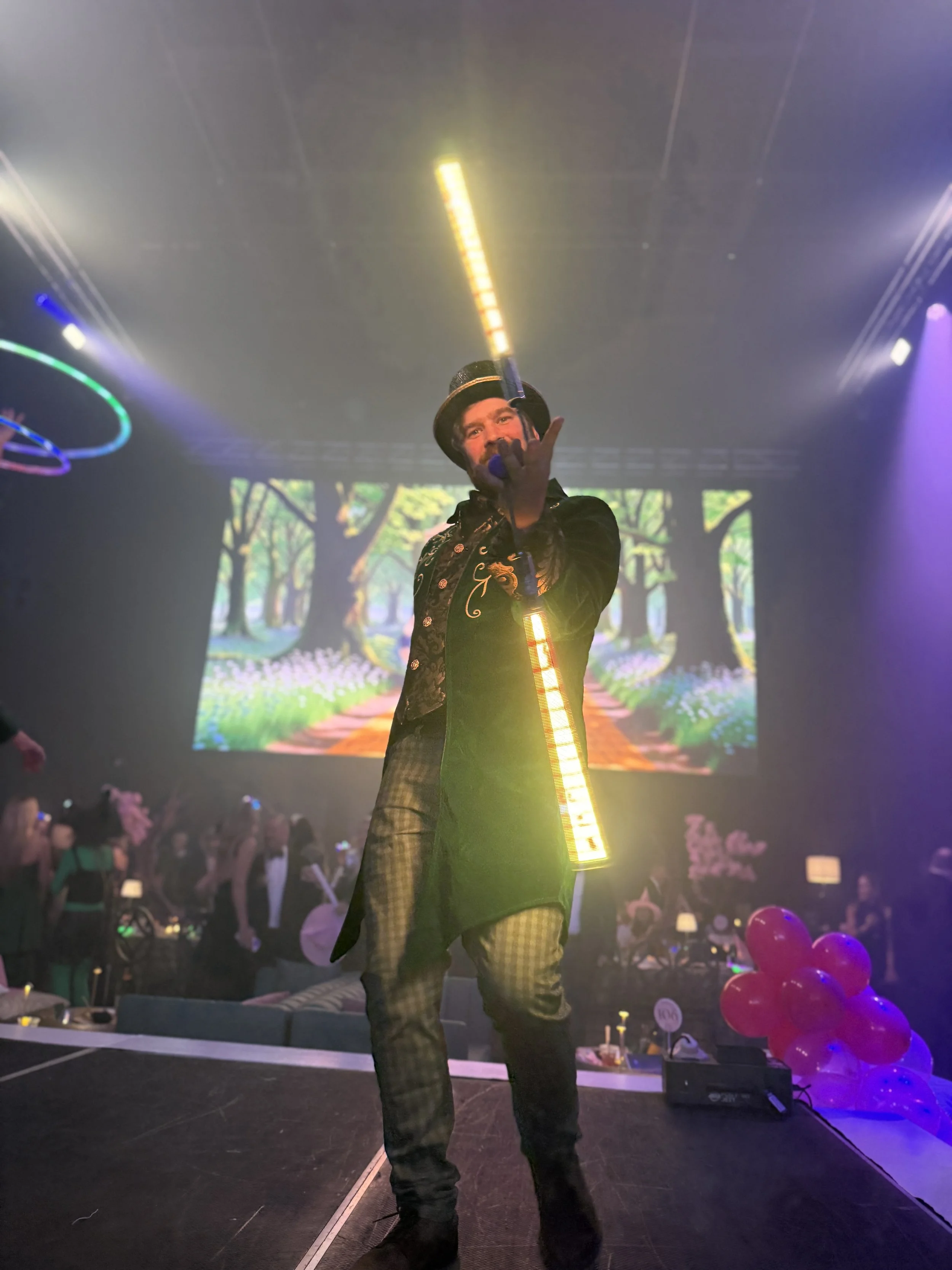 Performer on stage with LED lights and digital backdrop depicting a colorful forest path, audience in background, and pink balloons on the side.