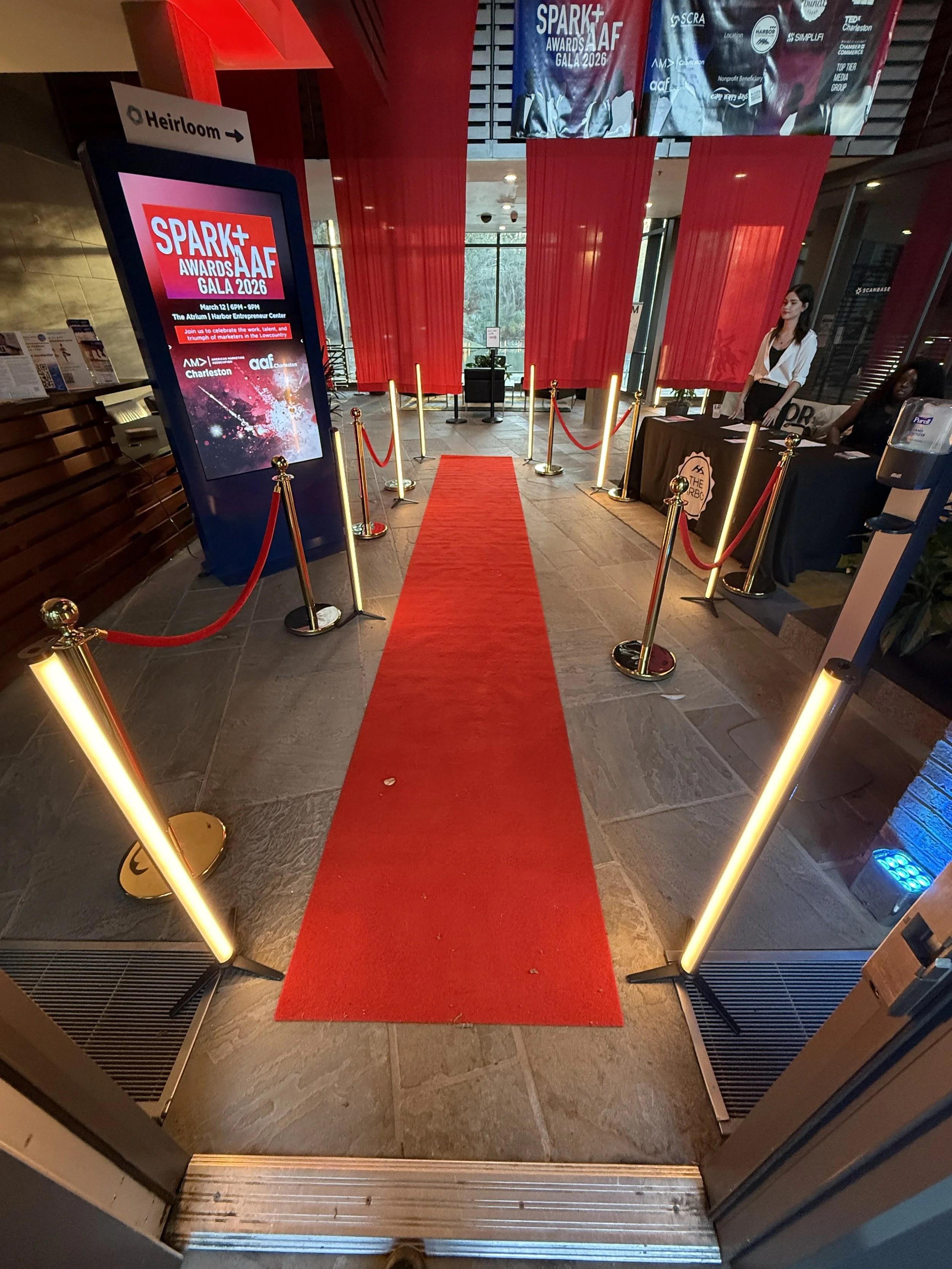 Red carpet leading to entrance of a conference or event at the Heirloom venue, with stanchions and ropes on both sides, digital sign on the left displaying details for the Spark+ Awards AAF Gala 2026, and registration tables on the right.