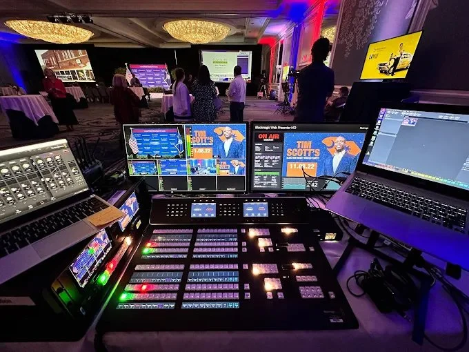 View of a conference or event with a stage and several screens displaying a news broadcast, in a decorated ballroom with chandeliers and people mingling.