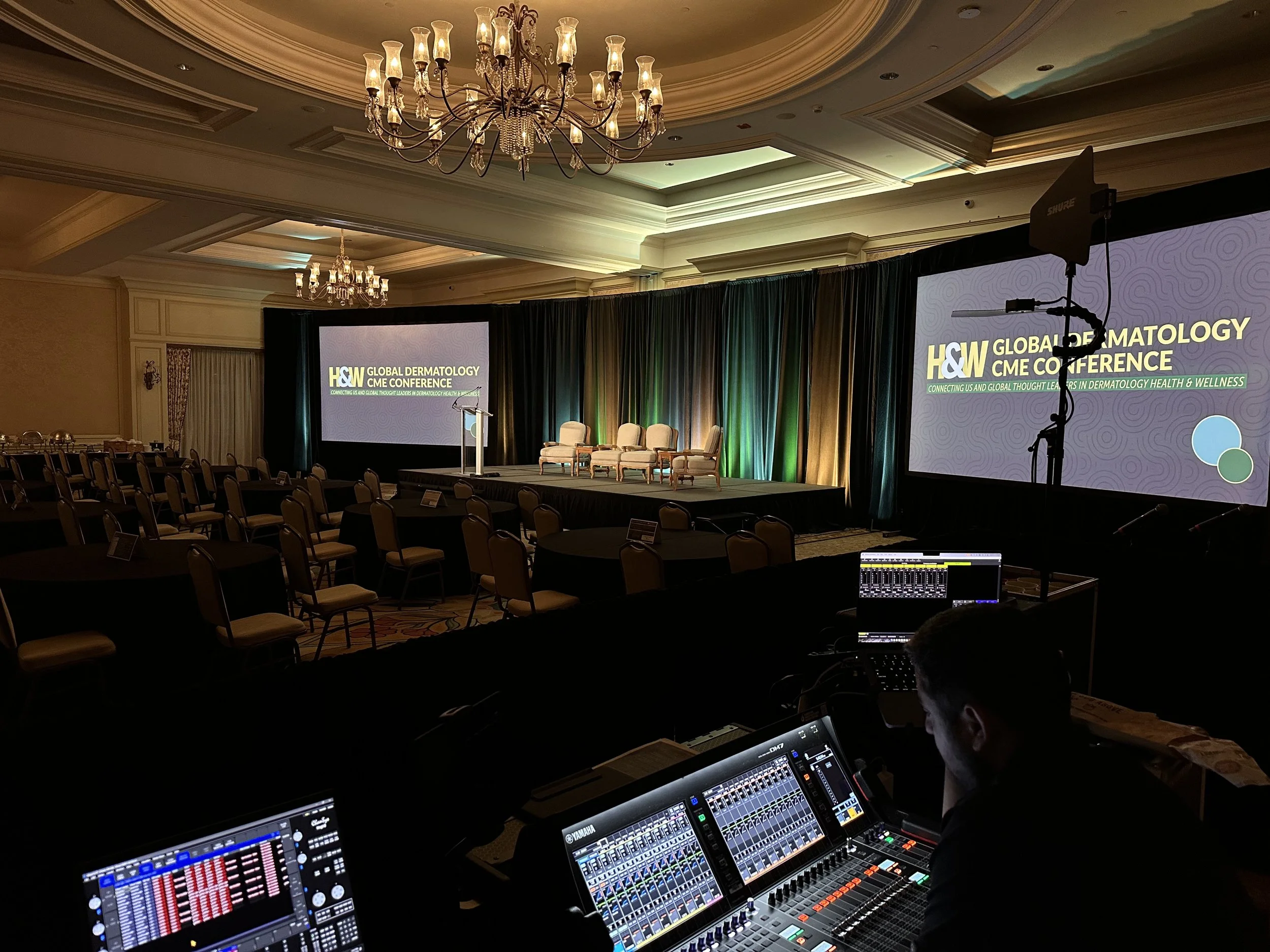 Conference stage set for the H&W Global Dermatology CME Conference with chairs and large screens displaying the conference title, and a person operating audio equipment in the foreground.