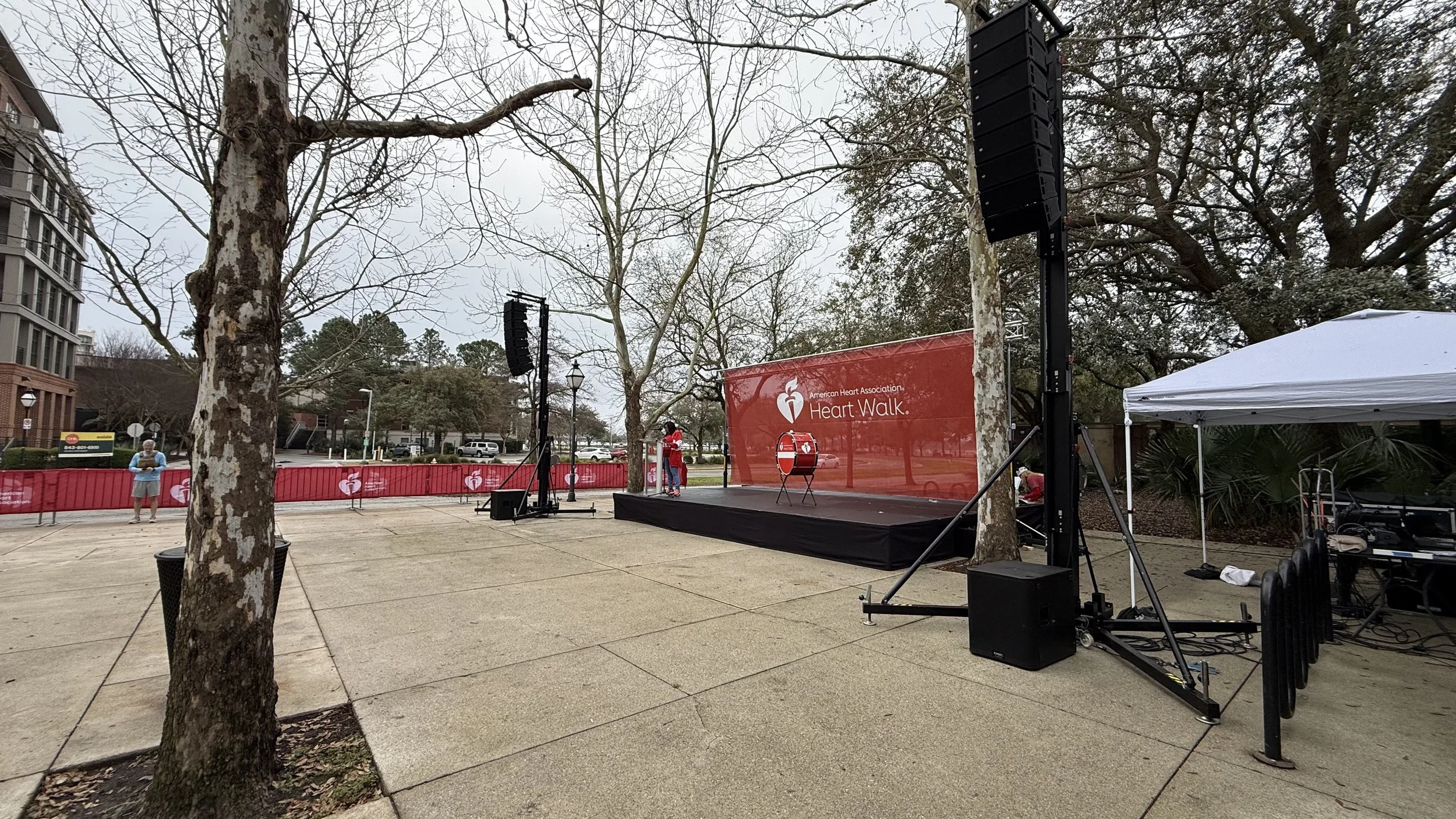 An outdoor stage set up for the American Heart Association Heart Walk, with a red backdrop and logos, speakers, a drum, and a few people preparing for the event on a cloudy day.