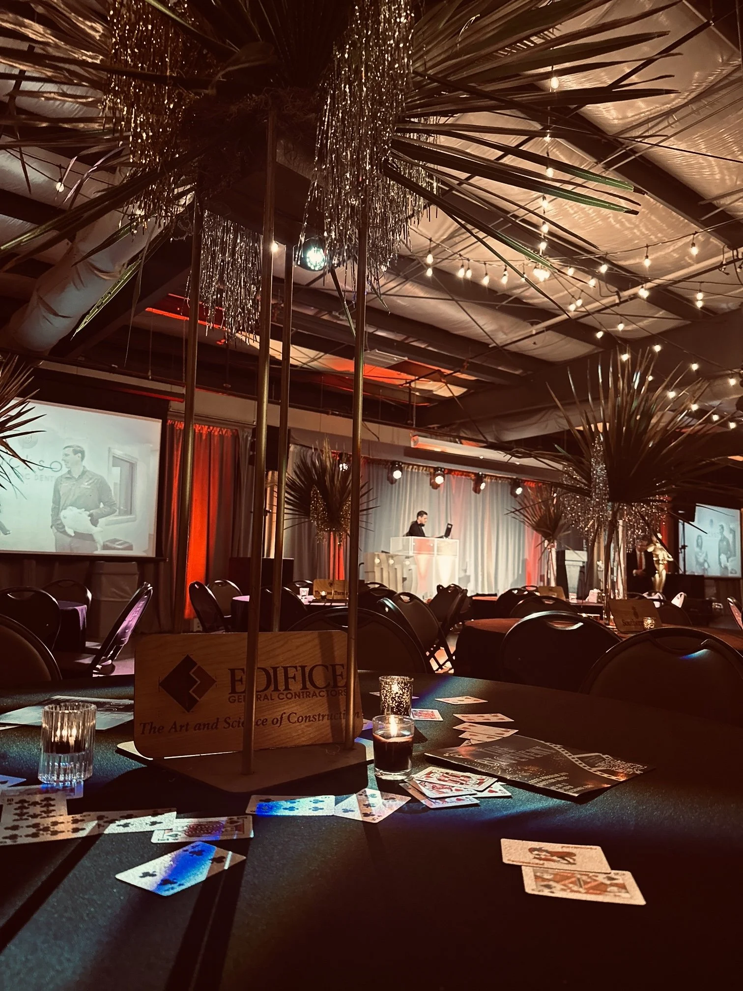 An indoor event with a stage and a DJ, decorated with string lights and tropical palm decorations, with tables featuring playing cards and candles.