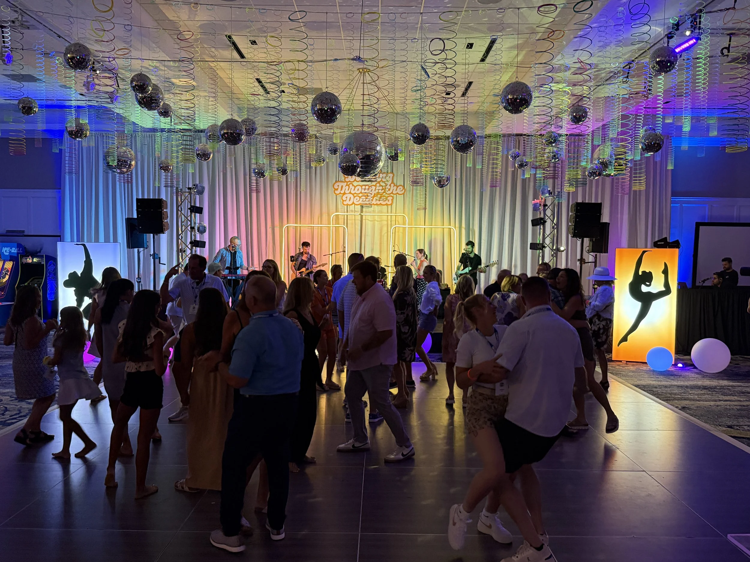 People dancing and enjoying live music at a vibrant indoor dance party with colorful lighting, disco balls, and dance floor decorations.