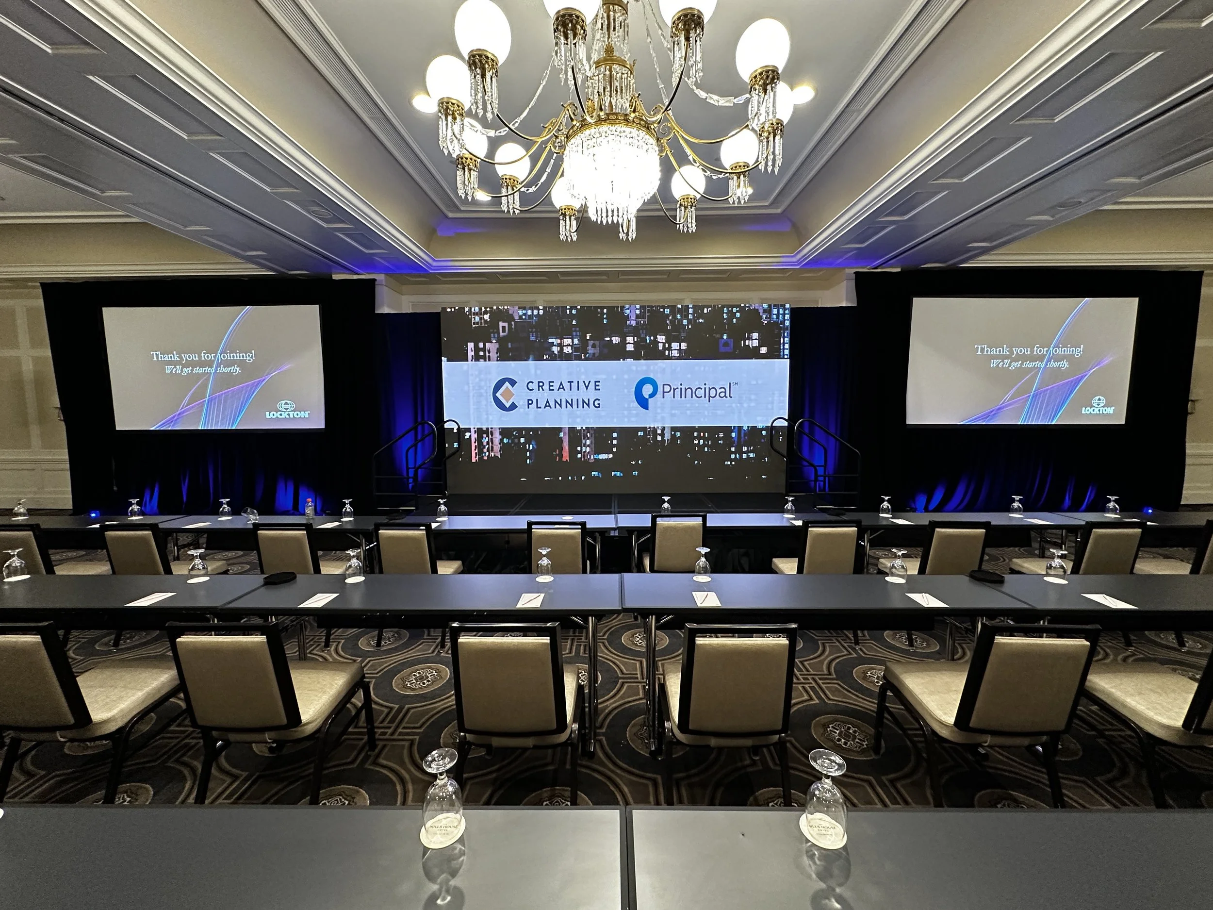 A large conference room with rows of tables and chairs, two projection screens displaying a message about a scheduled event, and a grand chandelier hanging from the ceiling.