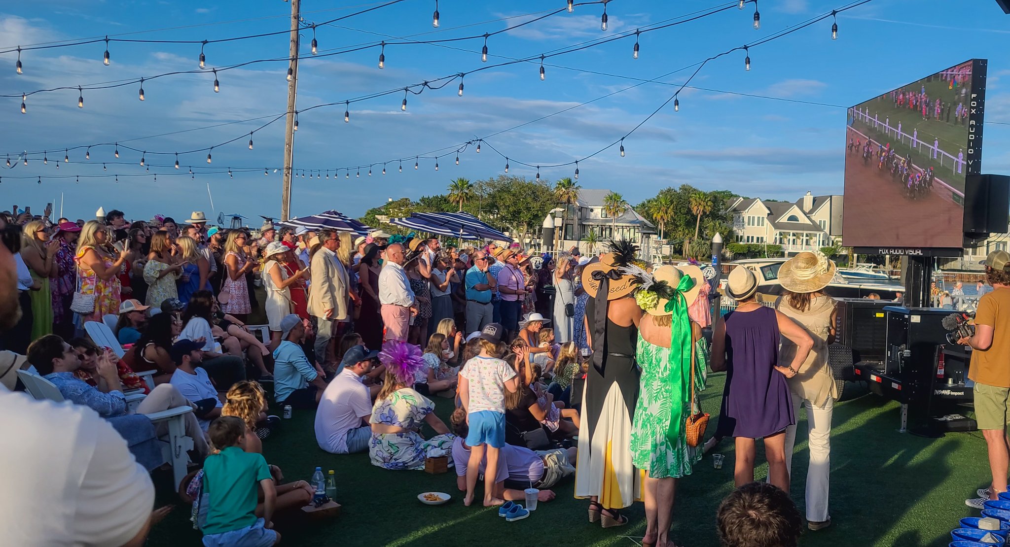 Crowd gathered outdoors watching horse racing on a large screen, with string lights overhead and people dressed in summer clothing and hats.