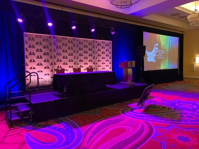 A stage set up for a presentation or conference with a podium, a table, and chairs. There is a large screen displaying a logo that says 'Fox Sports.' The stage is illuminated with purple and blue lighting, and the room has a patterned carpet.