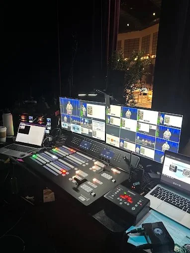 A video production control room with multiple monitors, a mixing console, and laptops.
