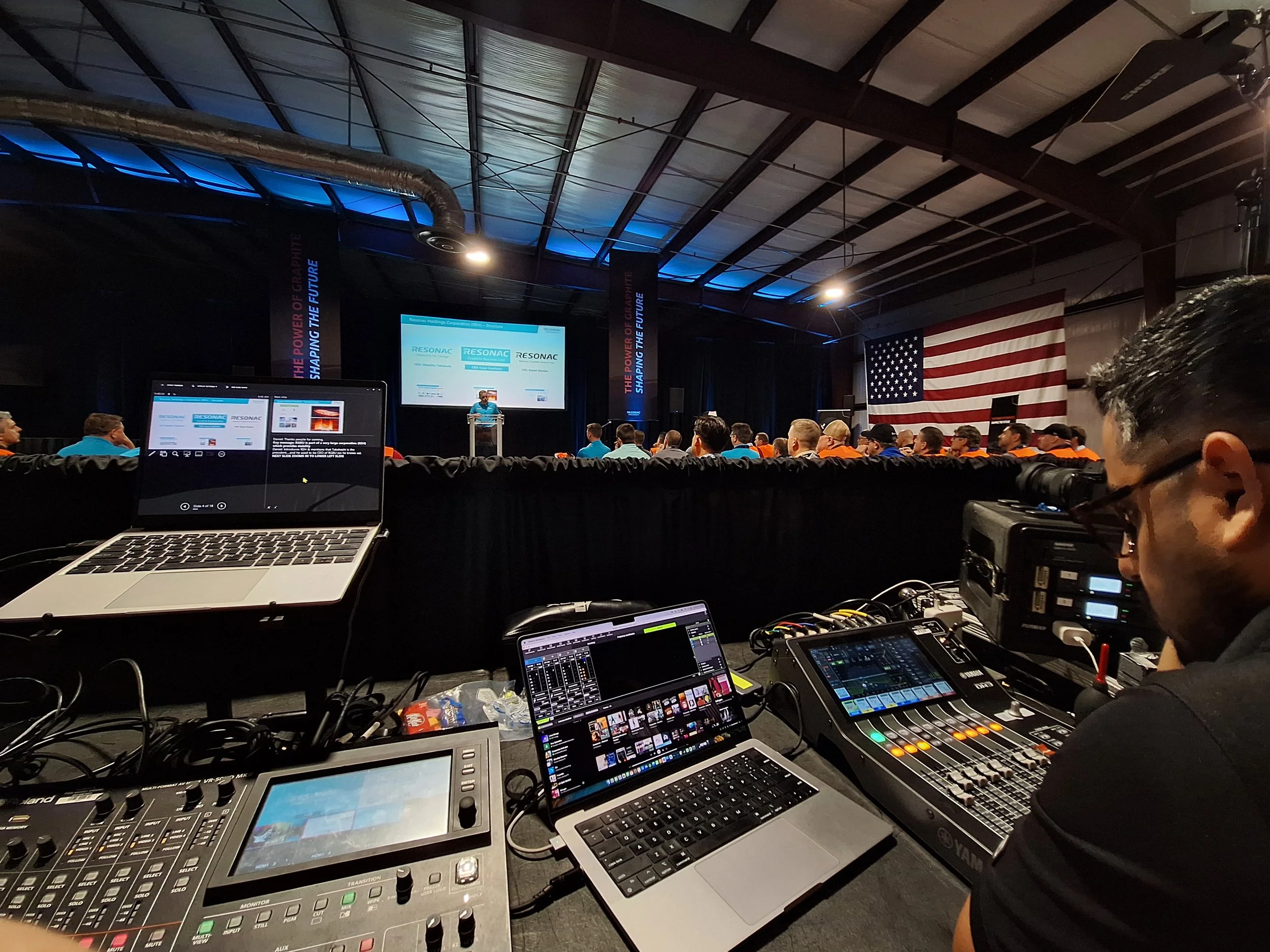 A large conference or presentation event with a stage showing a speaker at a podium and a large American flag hanging on the wall. Audience members are seated and a technician is working with audio and video equipment in the foreground.