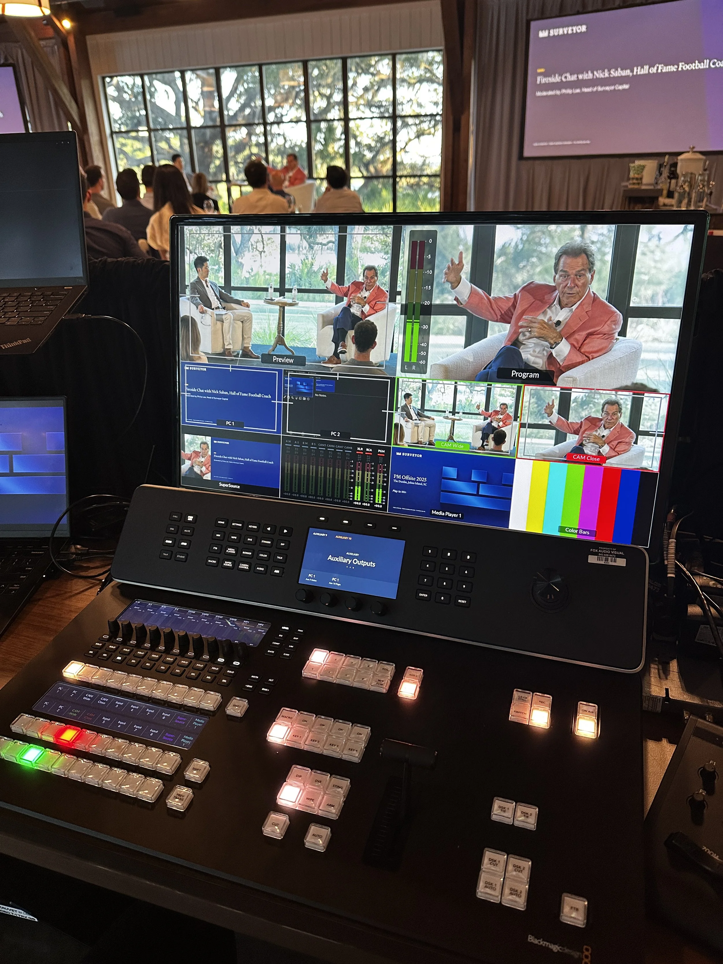 View of a broadcast control room with multiple monitors showing a panel discussion featuring two men seated in chairs, one wearing a red blazer. In the background, a large window with trees outside can be seen, and a conference room with people watch