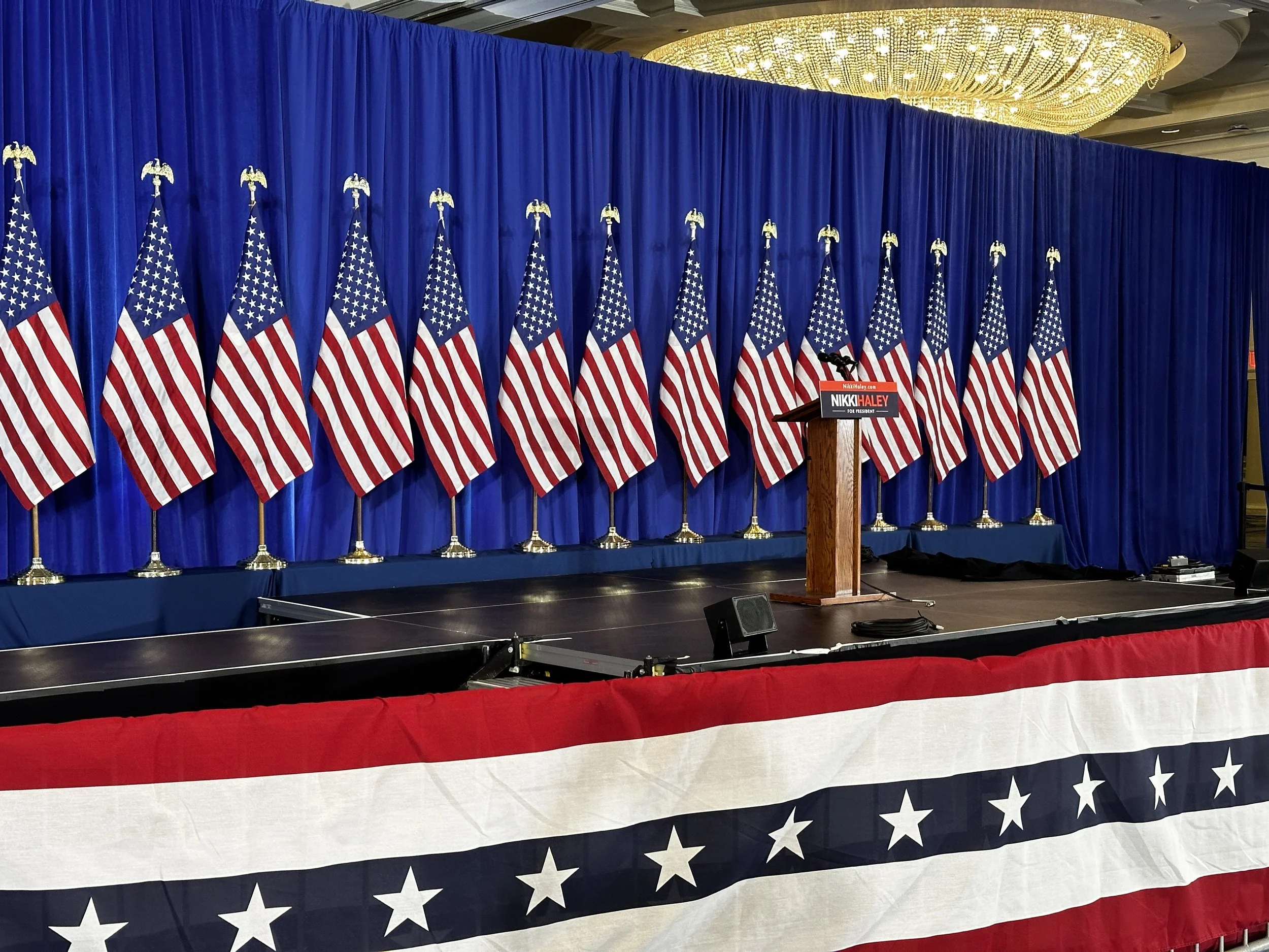 Multiple American flags are arranged in a row on a stage with a blue curtain backdrop. A podium with a sign reading 'Nikki Haley for President' is positioned at the right side of the stage. The ceiling has a large, ornate, golden chandelier, and ther
