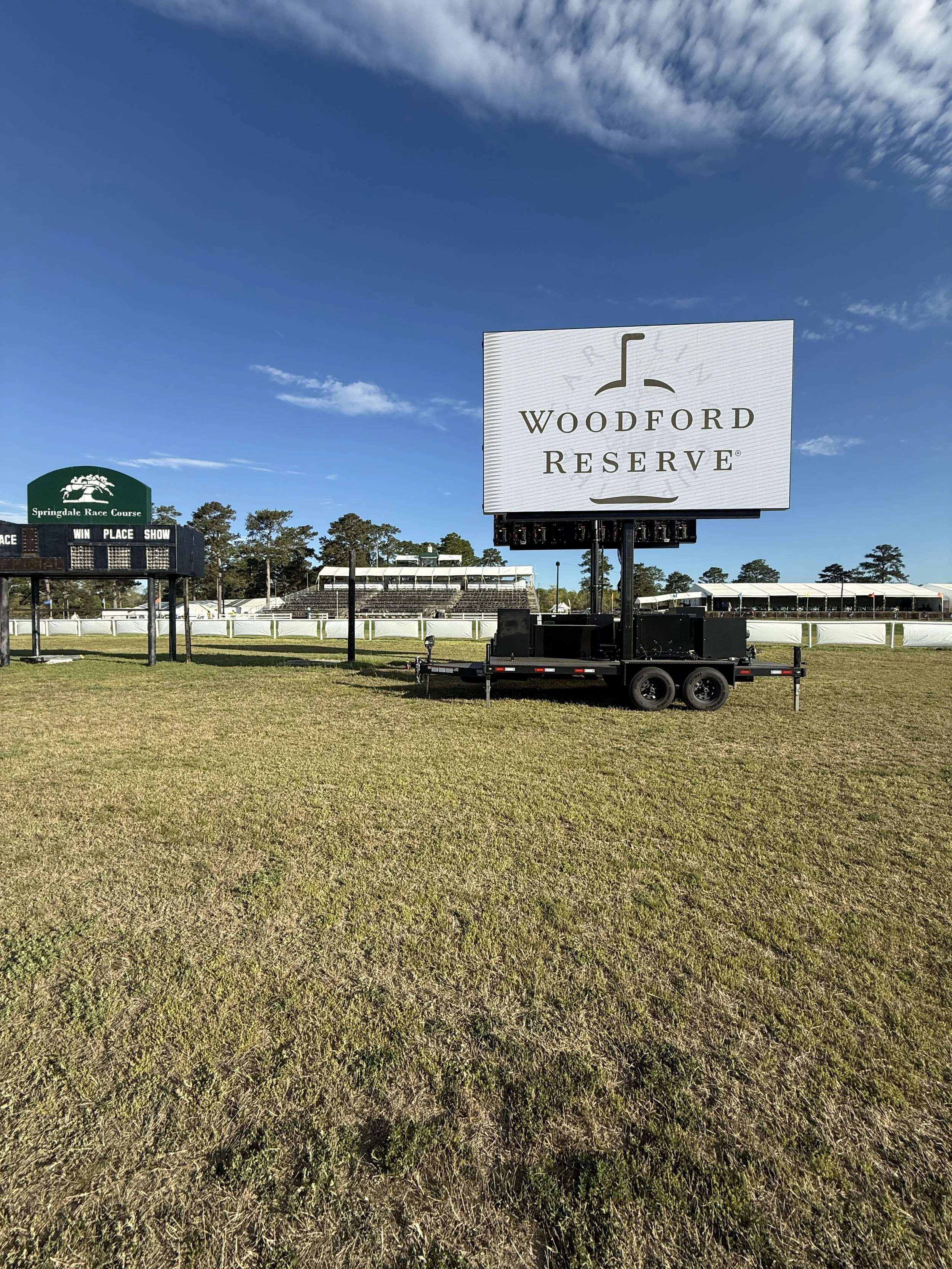 A large digital billboard at Woodford Reserve horse racing track displays the race branding with a partly cloudy sky overhead.