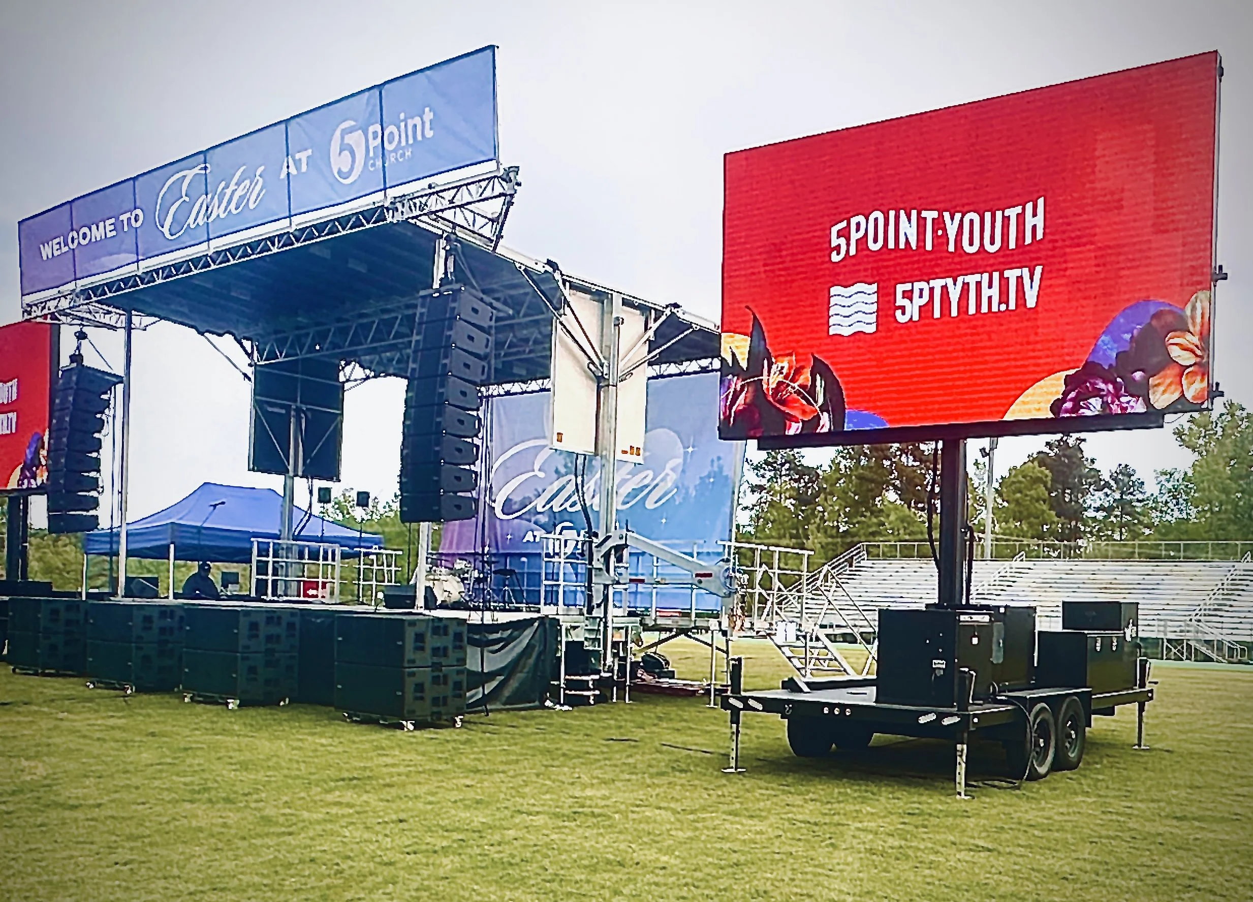 An outdoor stage setup with speakers, a large digital screen displaying '5POINT:YOUTH SPYTH.TV', and a canopy for shade. The stage is at Carter at 5 Point Church with empty bleachers in the background.