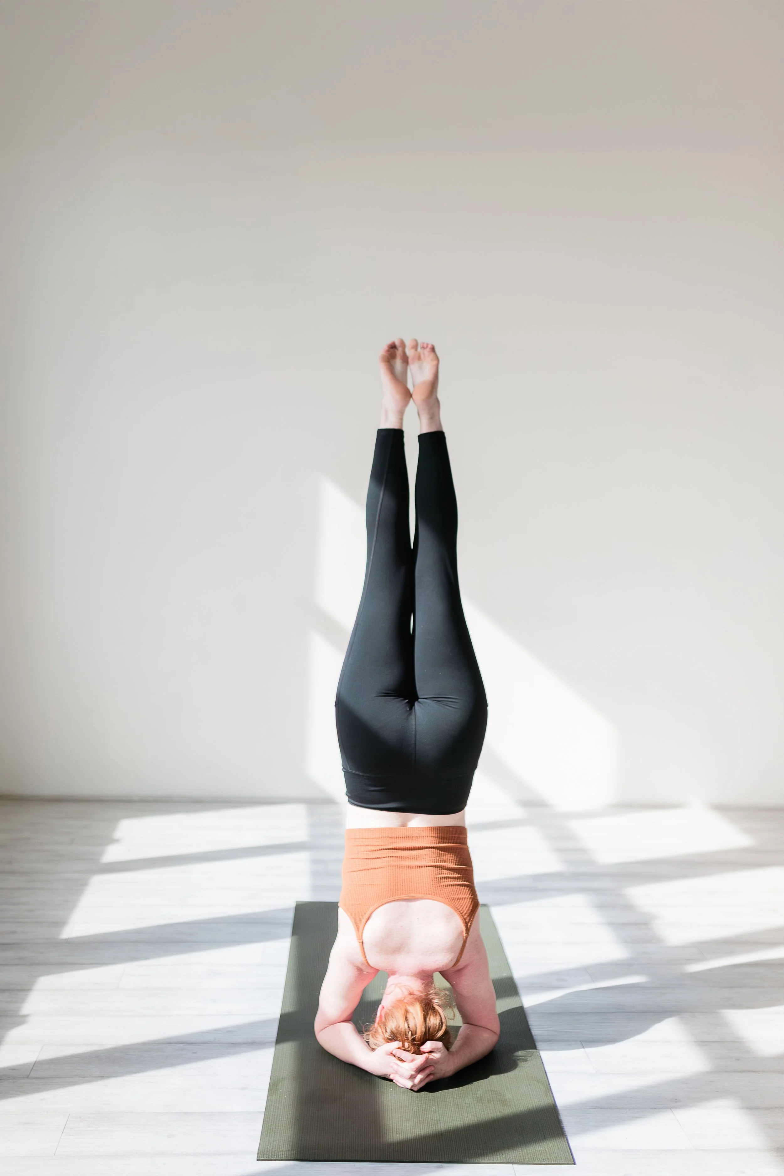 A woman with red hair practicing yoga in a headstand position indoors on a gray yoga mat, with a blank light-colored wall in the background and sunlight casting shadows on the floor.
