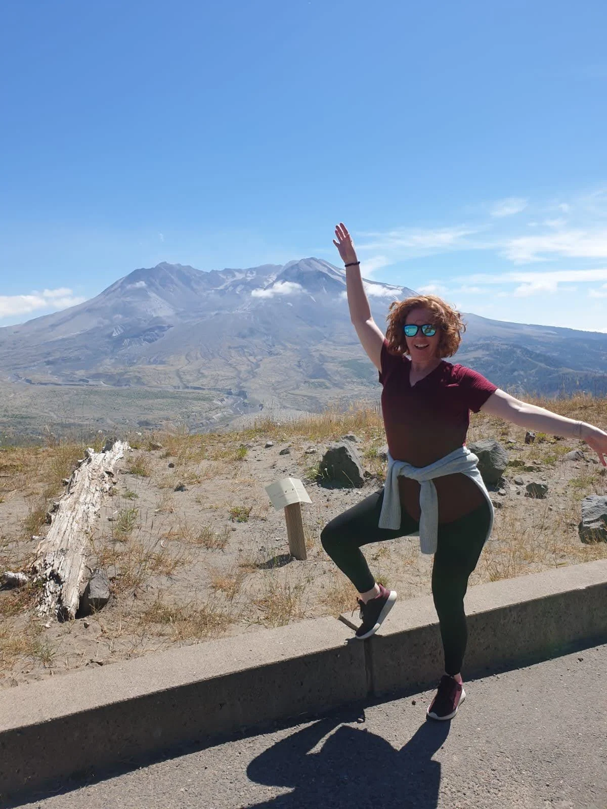 A woman with curly red hair, sunglasses, and a maroon shirt is posing playfully with arms outstretched and one foot on a curb in front of a mountain landscape, with a snow-capped volcano in the background under a blue sky.