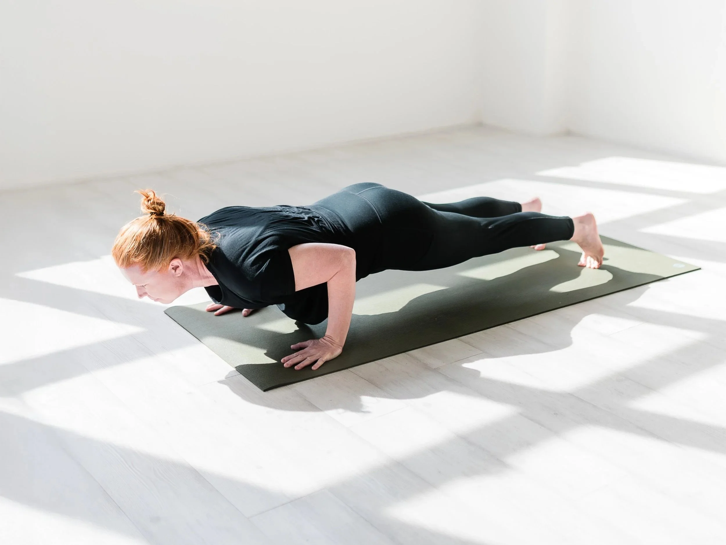 A woman with red hair practicing yoga in a plank position on a black mat in a bright, minimalistic room with white walls and natural sunlight.