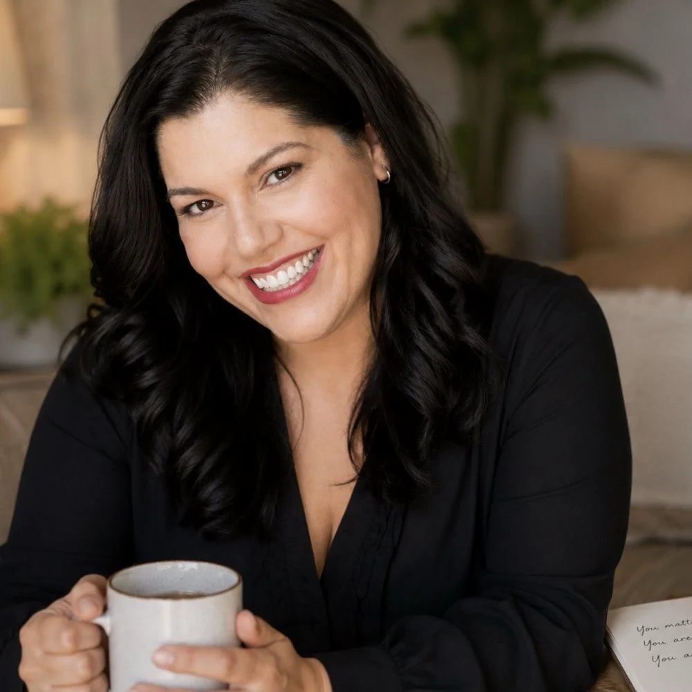 A woman with long black hair and a big smile holding a cup of coffee indoors.