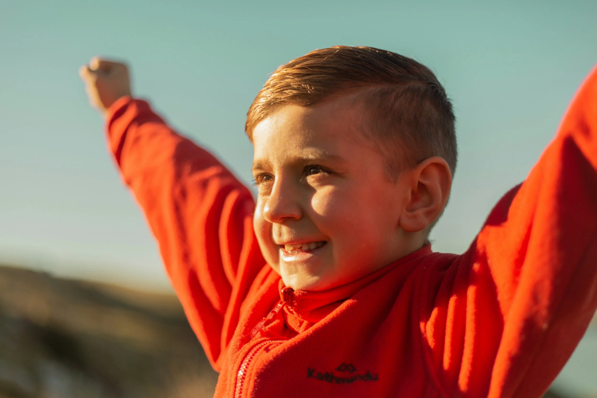 A young boy with short brown hair and bright eyes smiling broadly while raising his arms, wearing an orange jacket, outdoors against a blurred natural background.