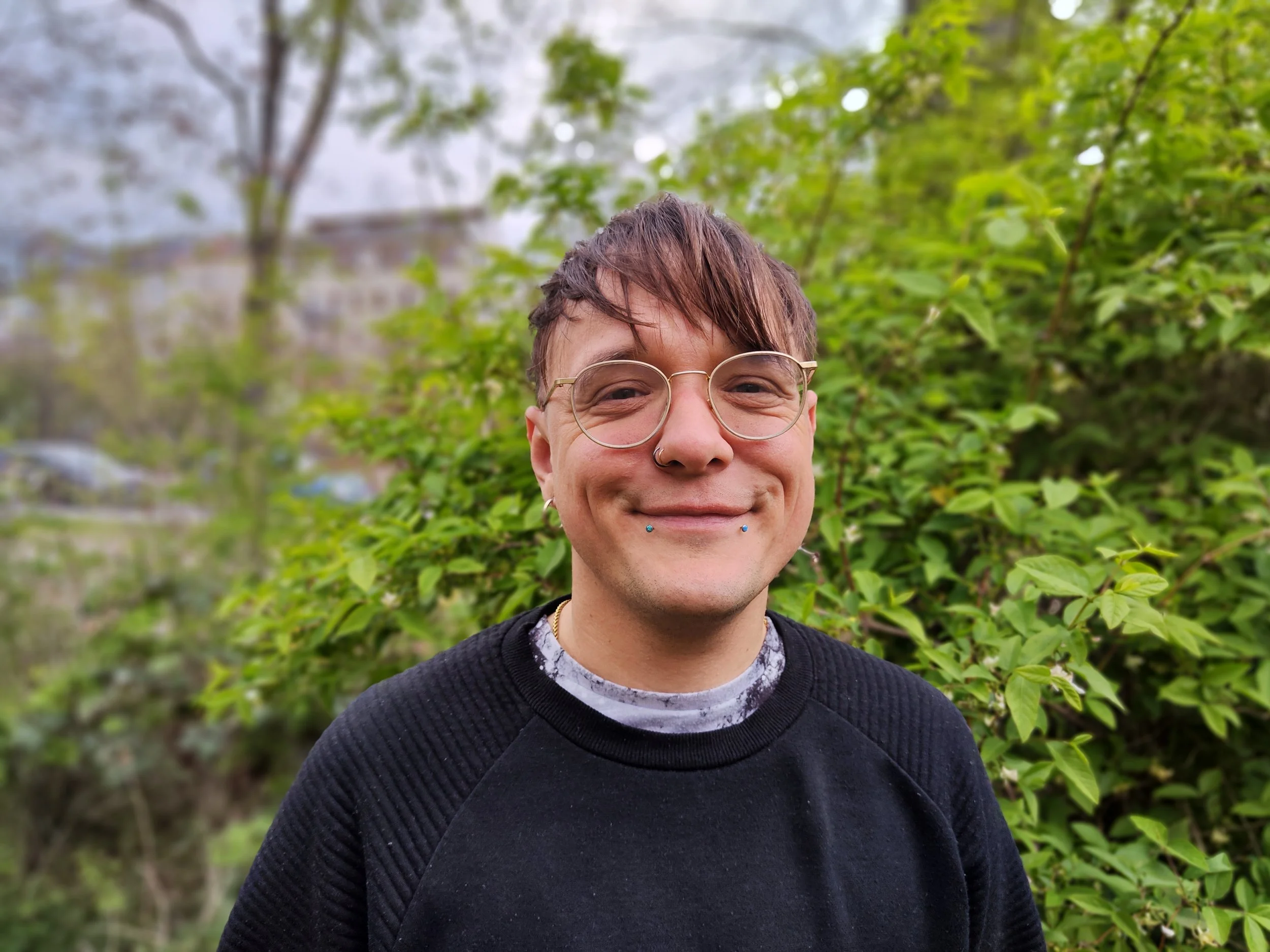 Nahaufnahme eines lächelnden Person mit kurzen braunen Haaren, Brille und Piercings im Gesicht, vor grünem Laub im Park.