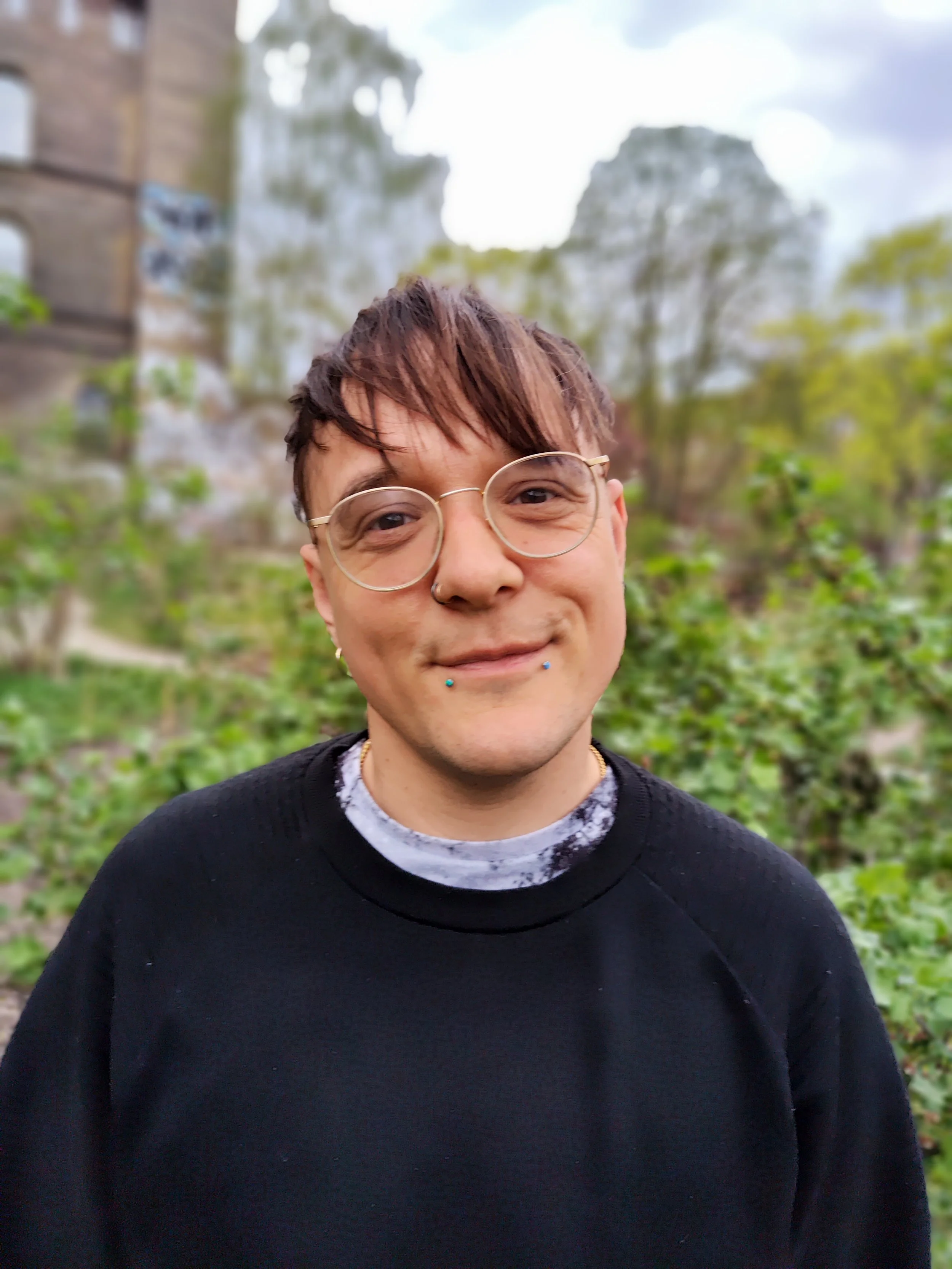 Nahaufnahme eines lächelnden jungen Mannes mit Brille, Piercings und dunklem Pullover vor einer grünen Baumkulisse mit einer alten Ruine im Hintergrund.