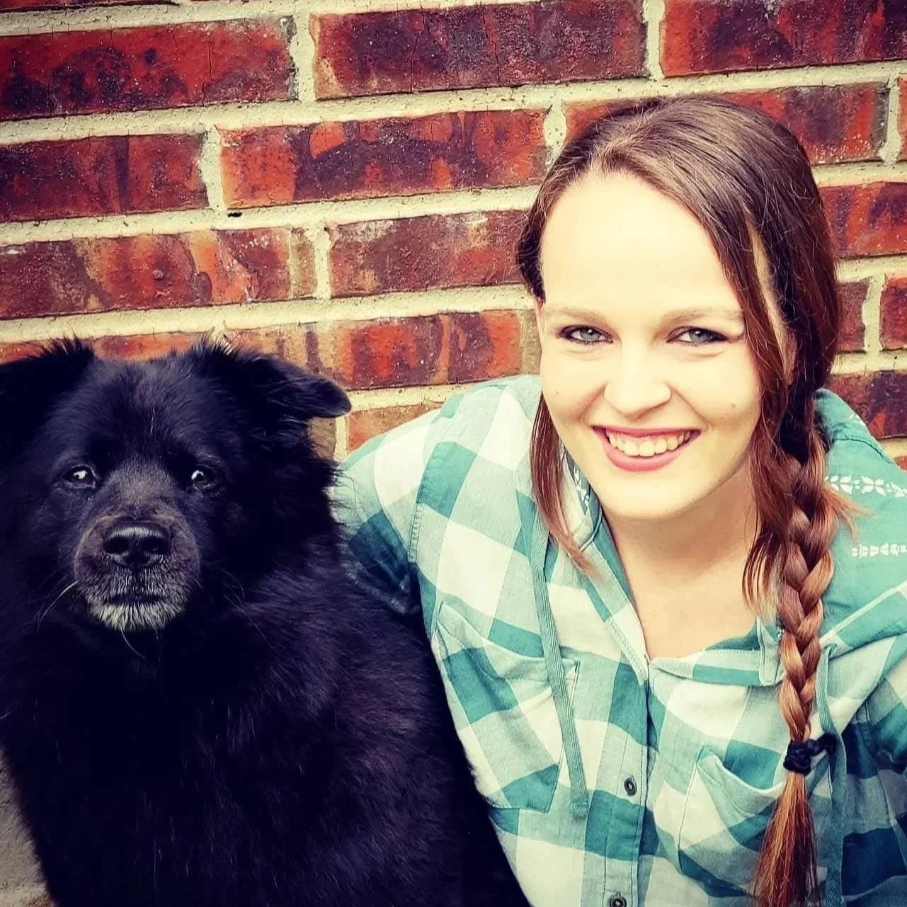 A young woman with long, reddish-brown hair in a braid, smiling, wearing a green and white checkered shirt, sitting next to a black dog with fluffy fur against a brick wall.
