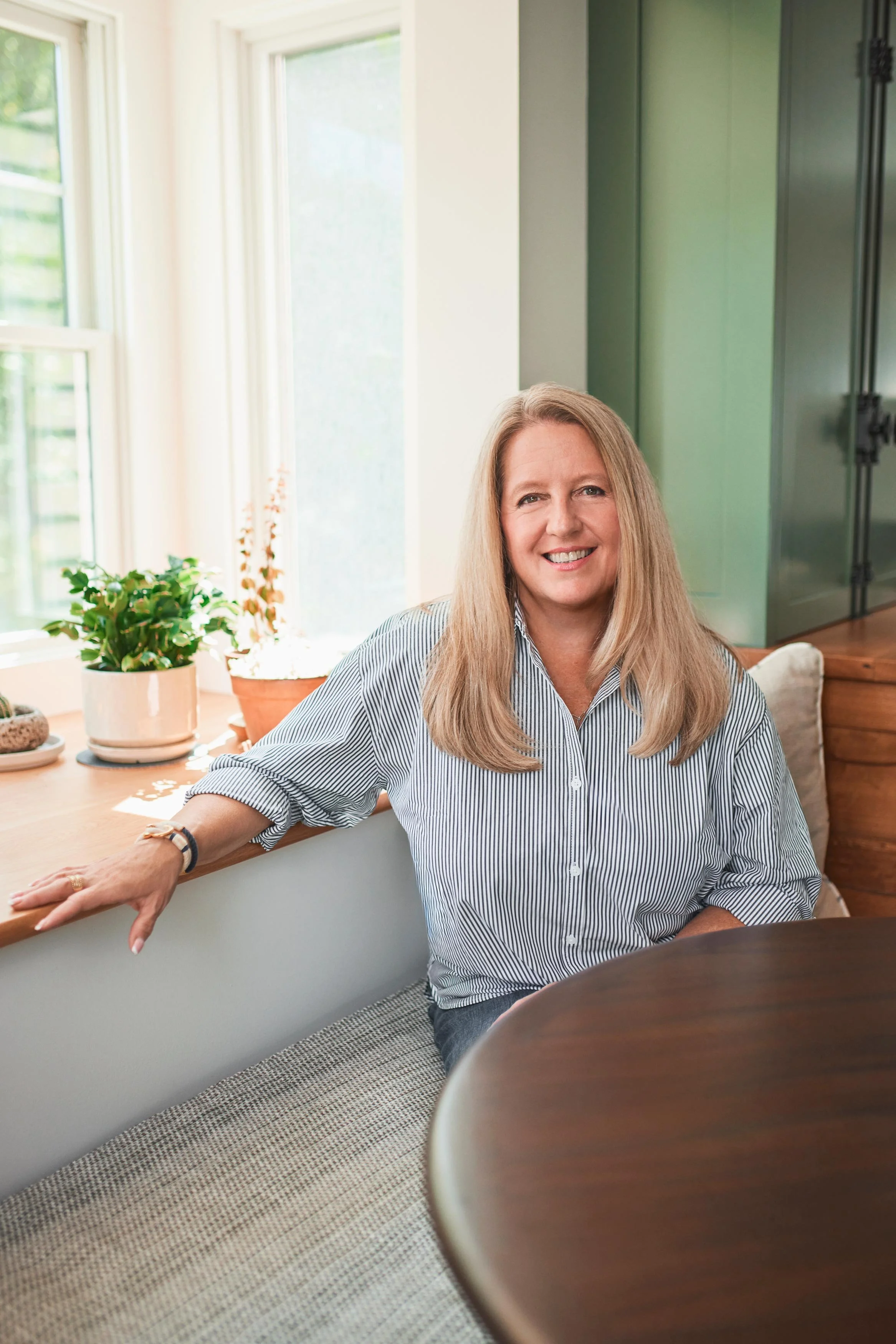 A smiling woman with long blonde hair sitting at a kitchen table, wearing a striped shirt, surrounded by potted plants near windows.