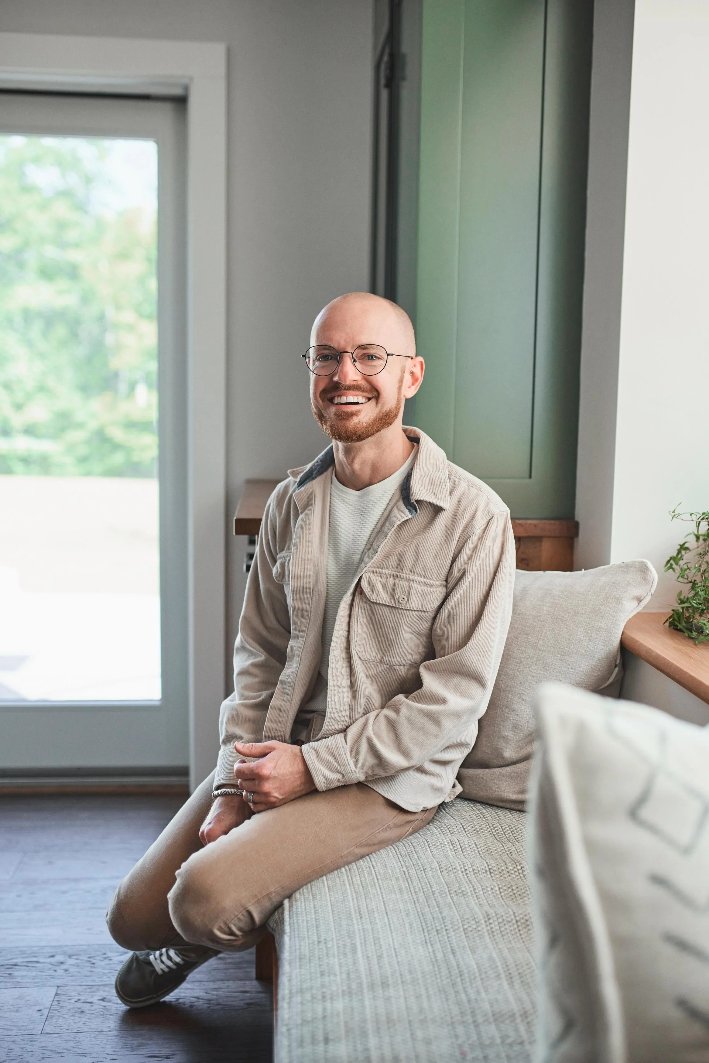 A man with glasses, a beard, and a bald head is sitting on a beige sofa in a bright, modern room. He is smiling and wearing a light-colored jacket over a white shirt, with his hands resting on his lap. Behind him, there is a green cabinet on the wall, a window showing greenery outside, and a plant on the right side of the image.