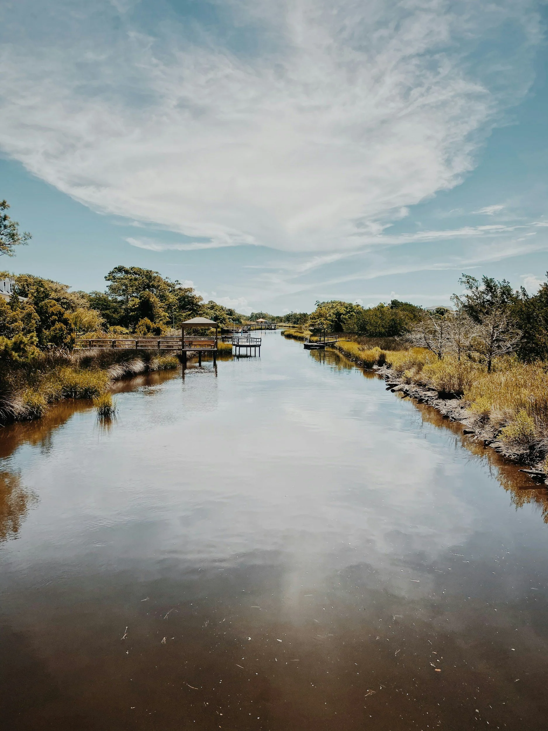 A calm waterway with docks and green trees along the riverbank under a cloudy sky.